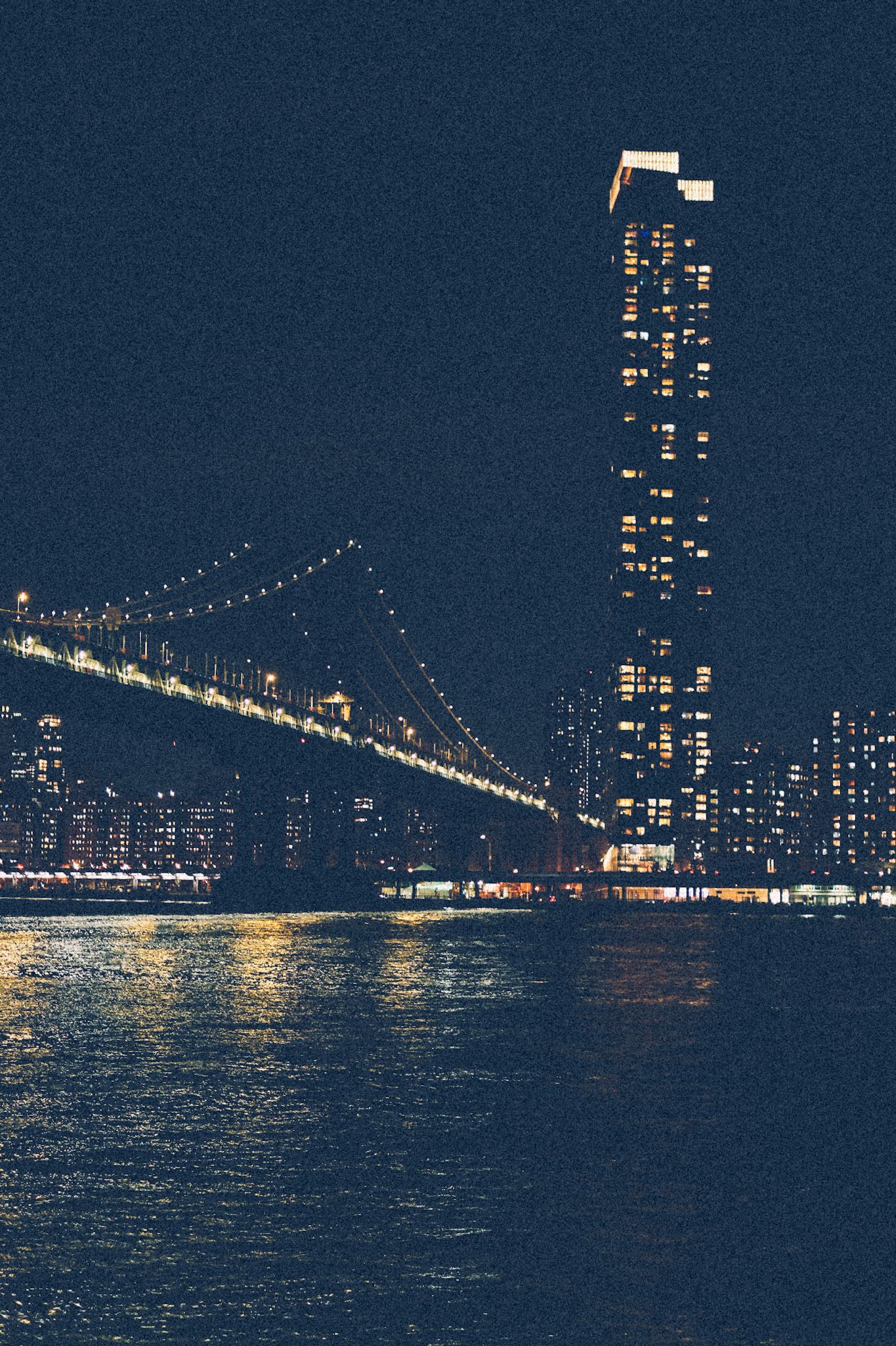 A view of the Manhattan bridge from downtown Brooklyn, overlooking a skyscraper at night. City lights sparkle.