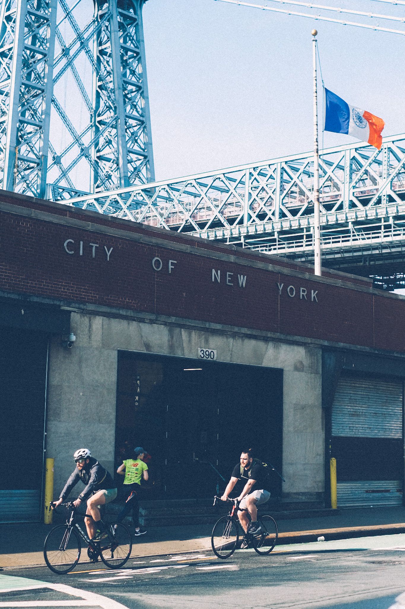 Two cyclists and a runner pass in front of a short brick building with “City of New York” on the outside of it. A blue, white, and orange flag flies on top of the building and a bridge is in the background. The sun is out.