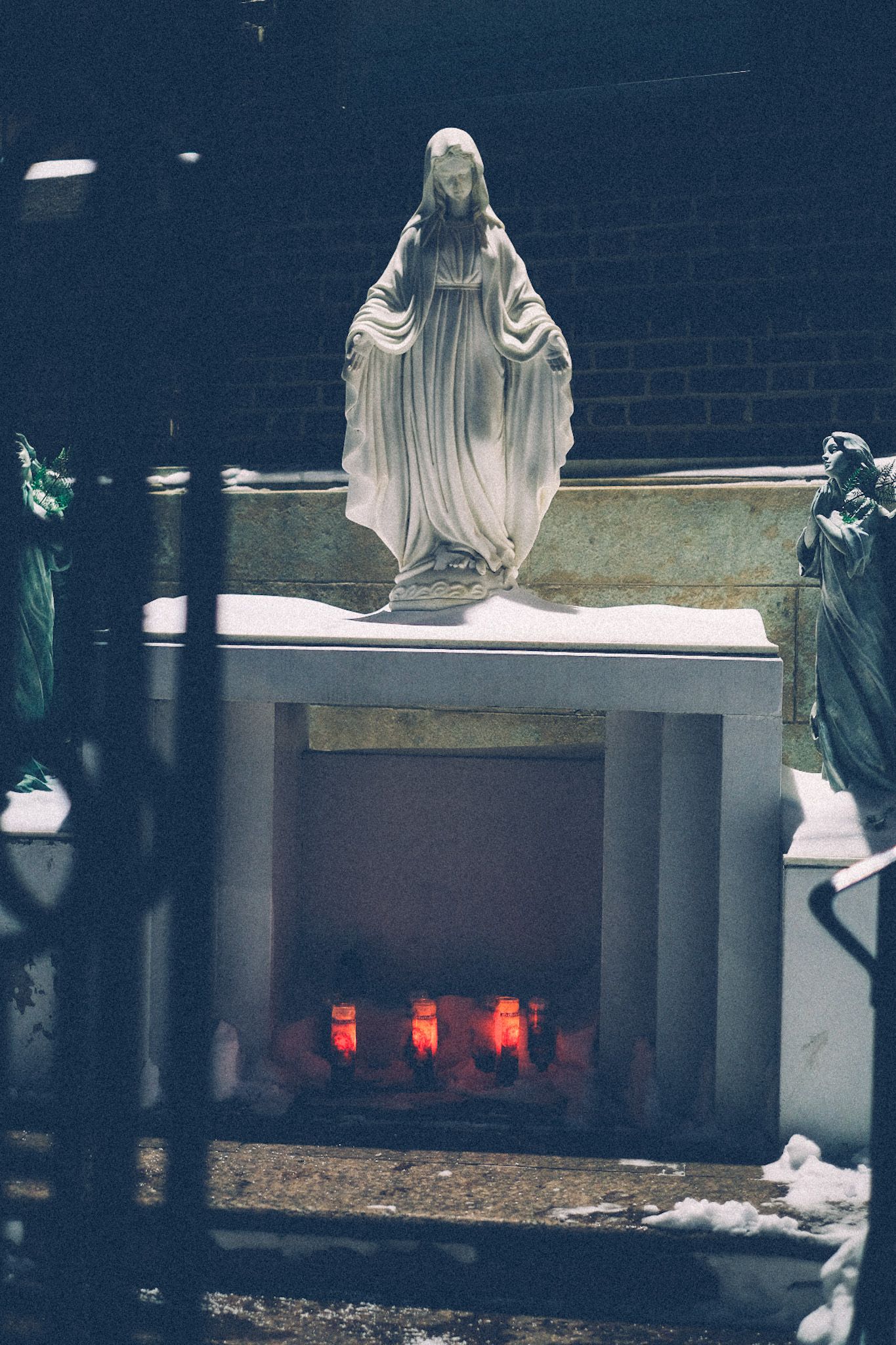 A statue of Mary sits atop a small alter housing red vigil candles, flickering in the evening. Two angels flank her sides under the light outside the brick church.