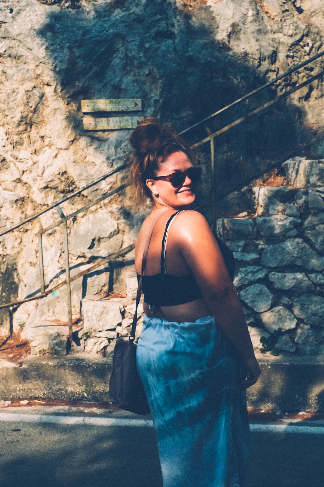 A woman in sunglasses with her hair in a bun looks over her shoulder at the camera. You can’t see her eyes because of the sunglasses. A jagged shadow shows on a cobblestone wall behind her, golden hour.