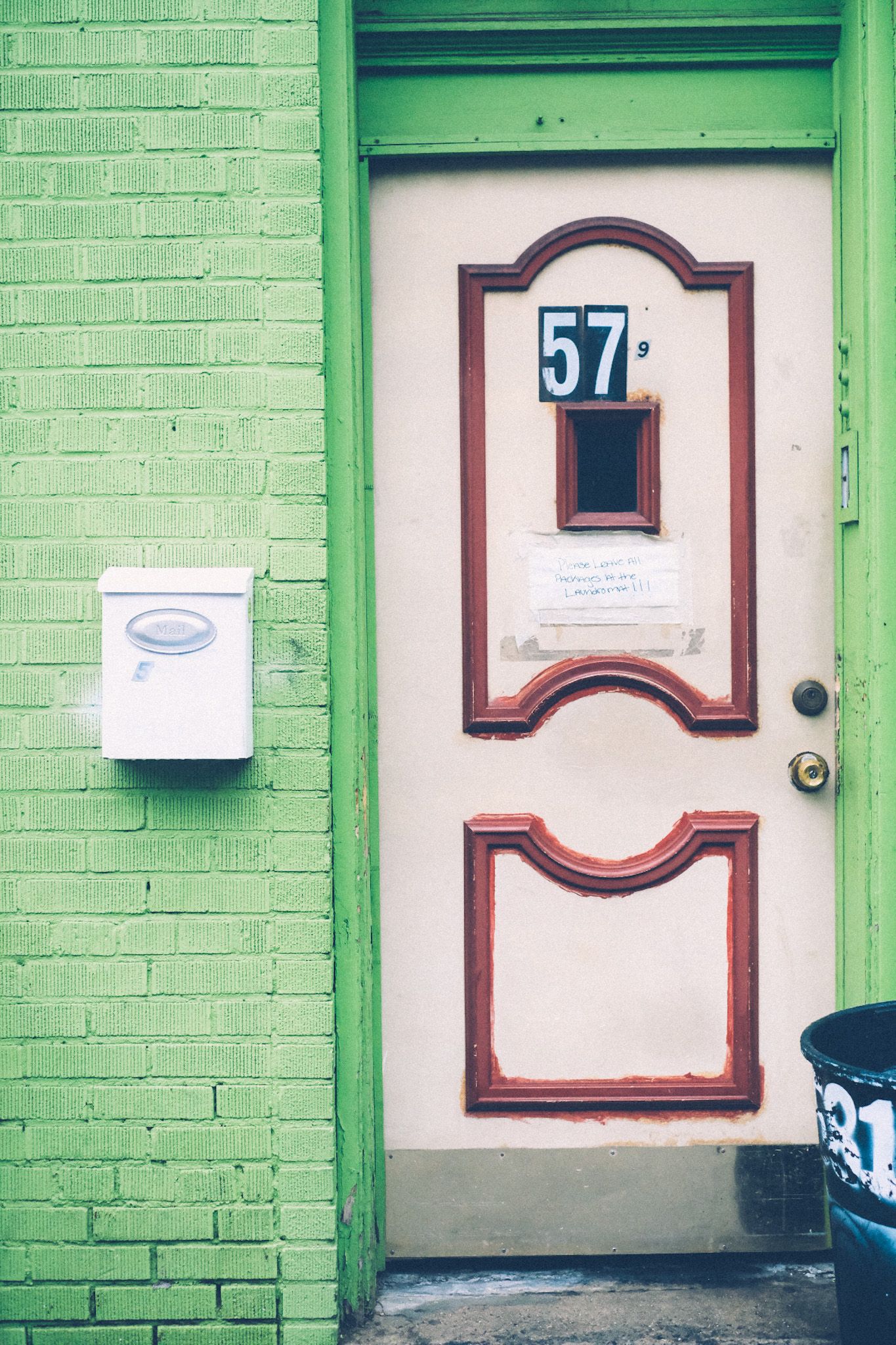 A lime green brick wall frames a cream white door with red detailing, the number 57 stuck on the front of the door along with a handwritten note saying “Please leave all packages at the laundromat!!!”. A white mailbox hangs on the wall to the left, and the corner of a black trash can peeks out on the right.