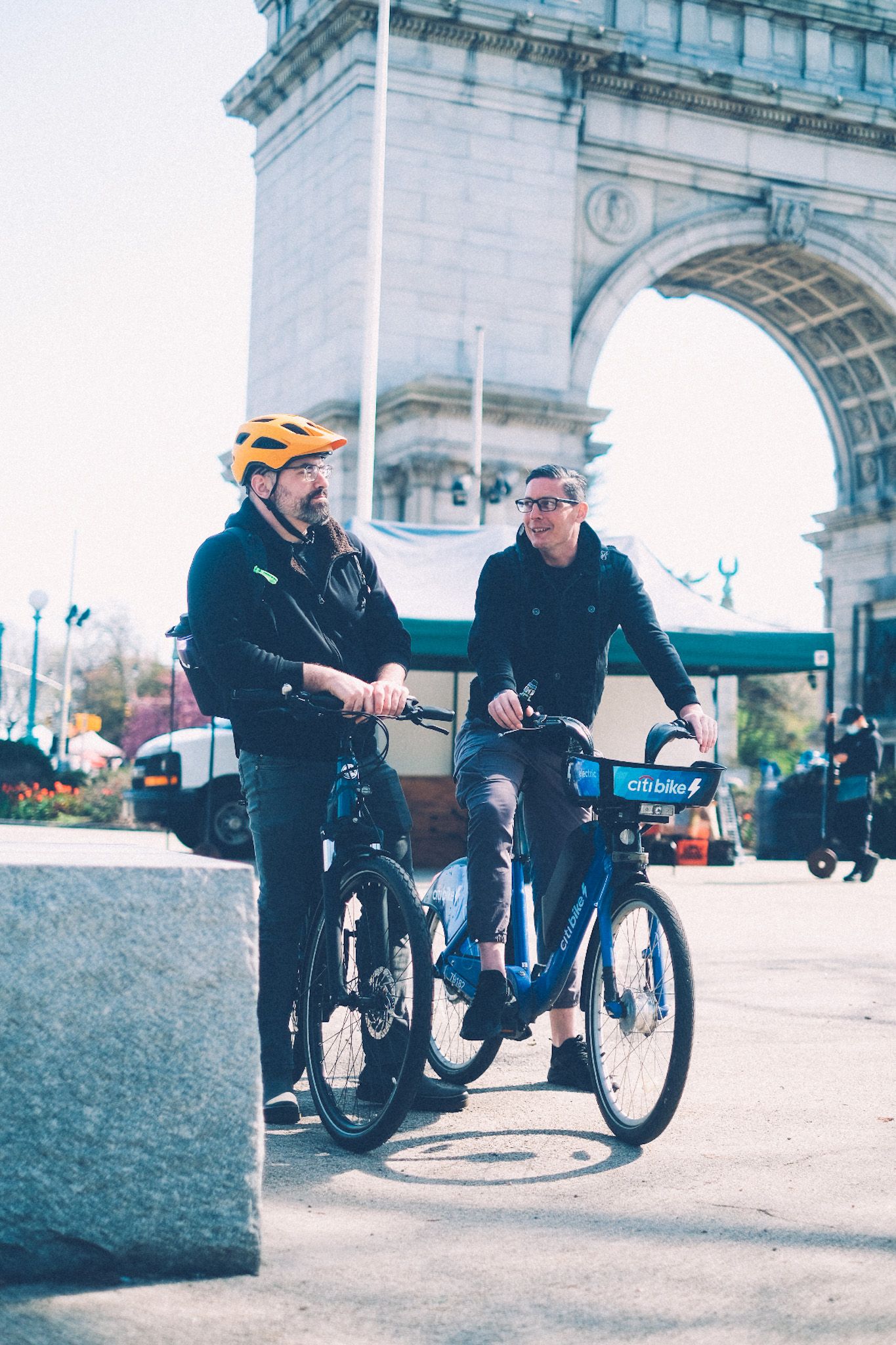 Two men are perched on their bikes in front of an archway outside of a park, talking to each other.