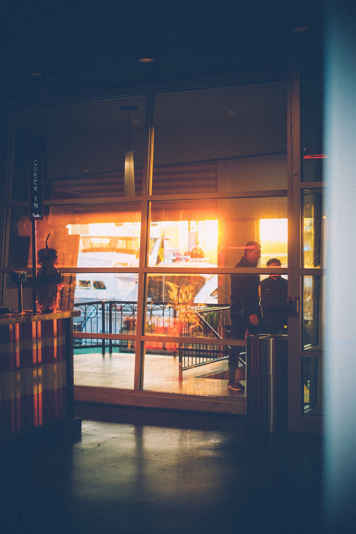 People pass in front of a glass window, where the sunset shines through into the entrance of a building that looks out onto the harbor.