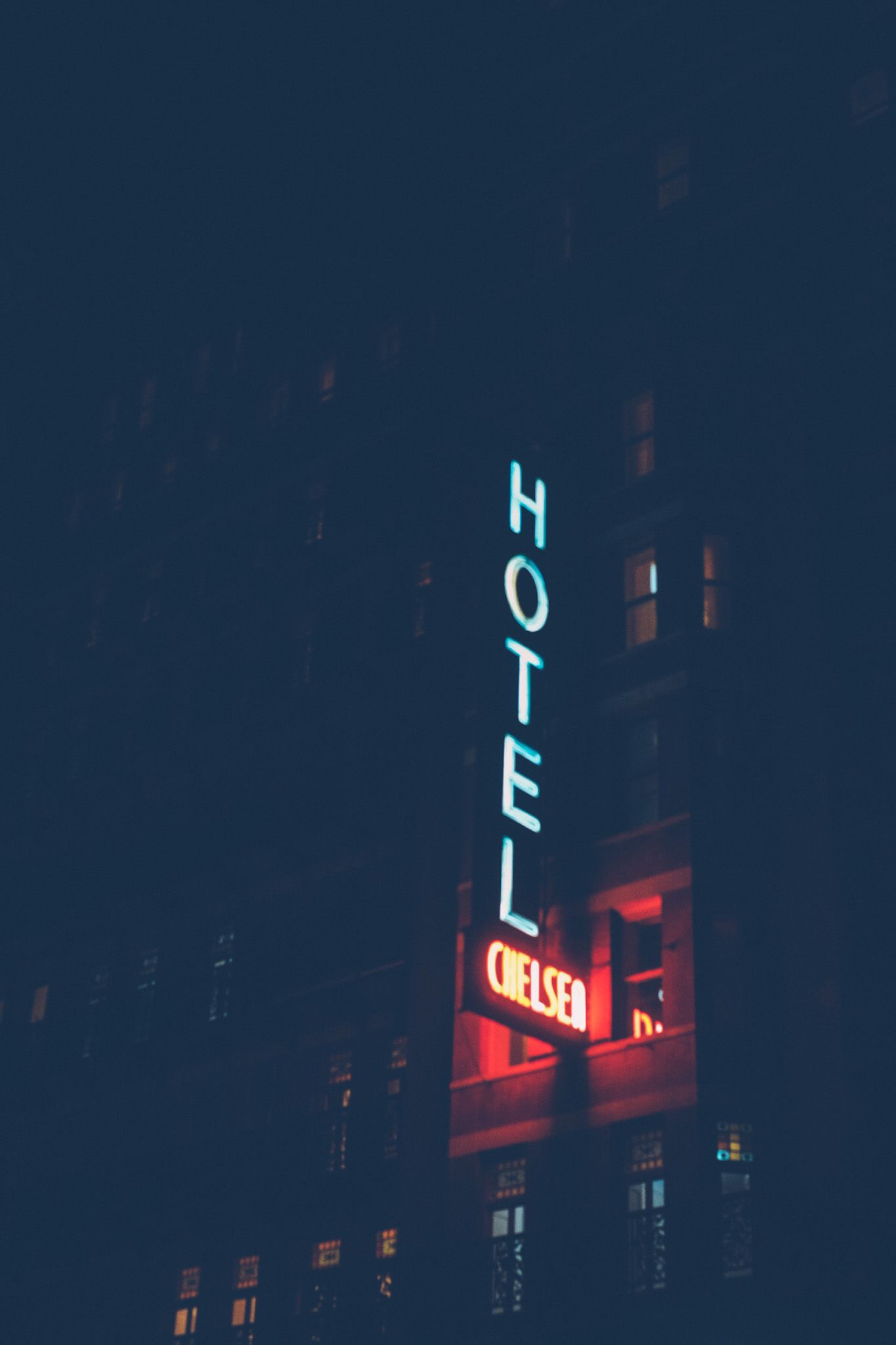 A neon sign saying “Hotel Chelsea” glows in the night, apartment windows lit behind it.