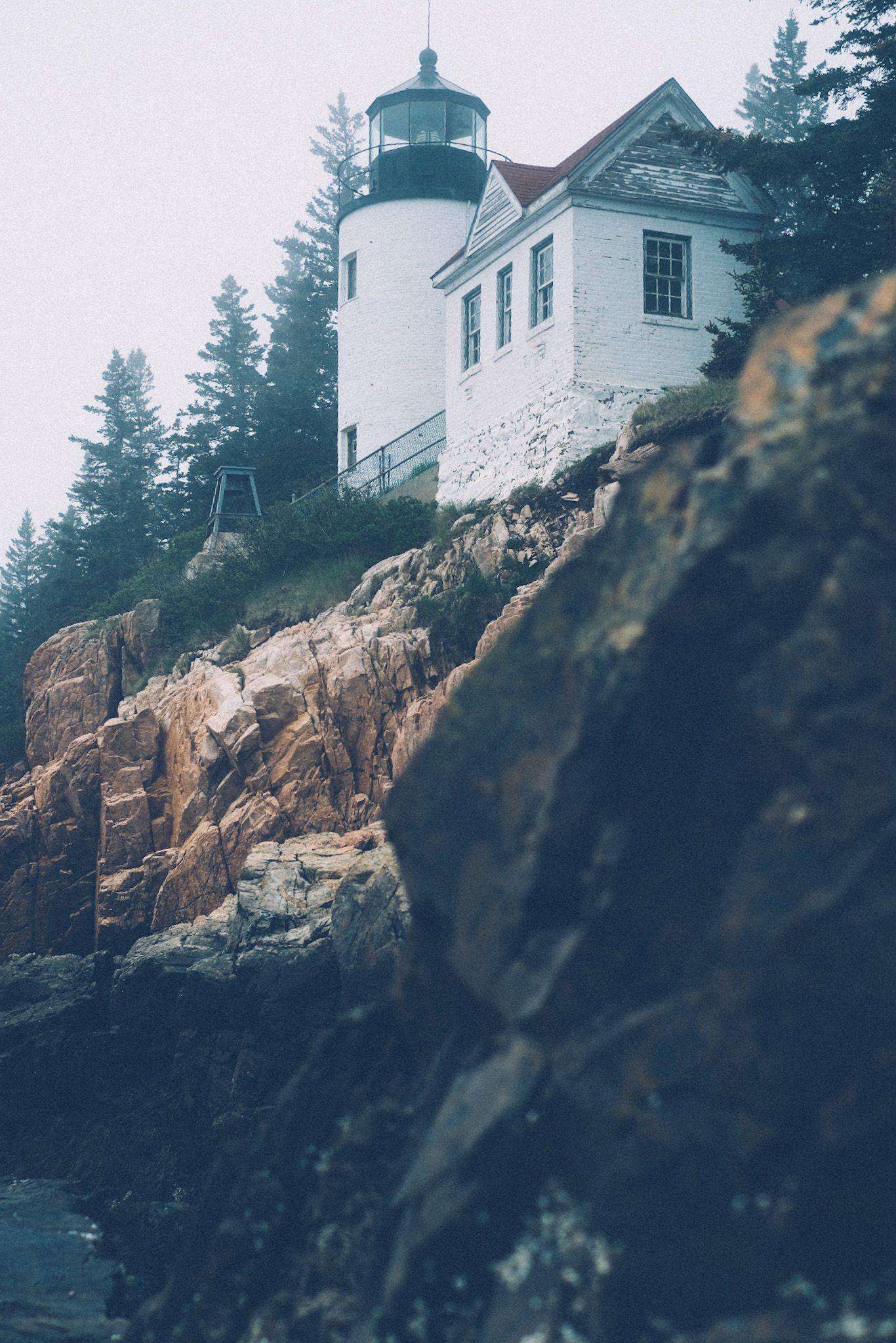 A lighthouse sits atop a foggy cliff, surrounded by sand-colored and dark black rocks beneath it.