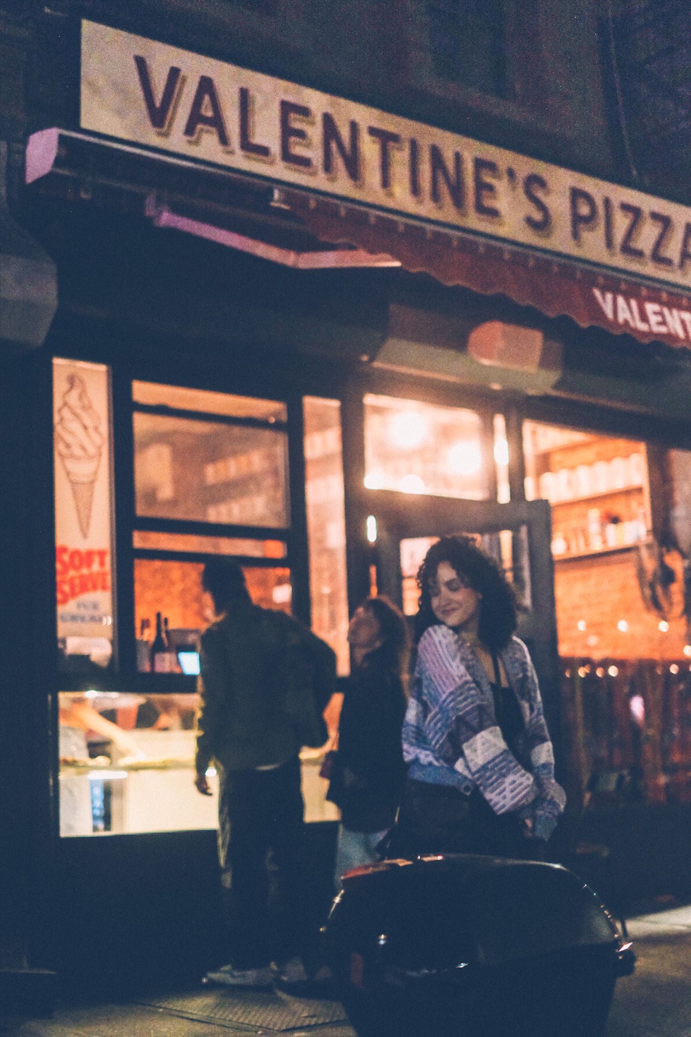A woman stands mid-ground in front of a storefront that says Valentine’s Pizza, at night, while people stand at the window ordering pizza behind her, just out of focus.