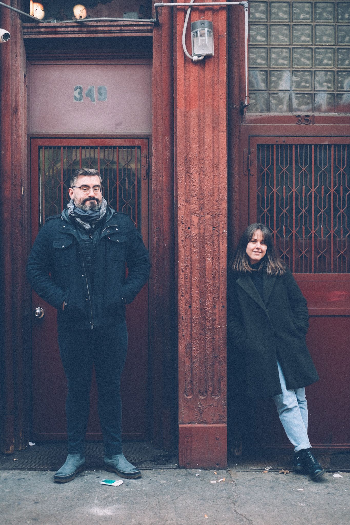 A man and a woman each stand in front of a rusty red door of an old building, split by a pole. They wear winter coats. The pattern on the doors is elegant and squiggled.