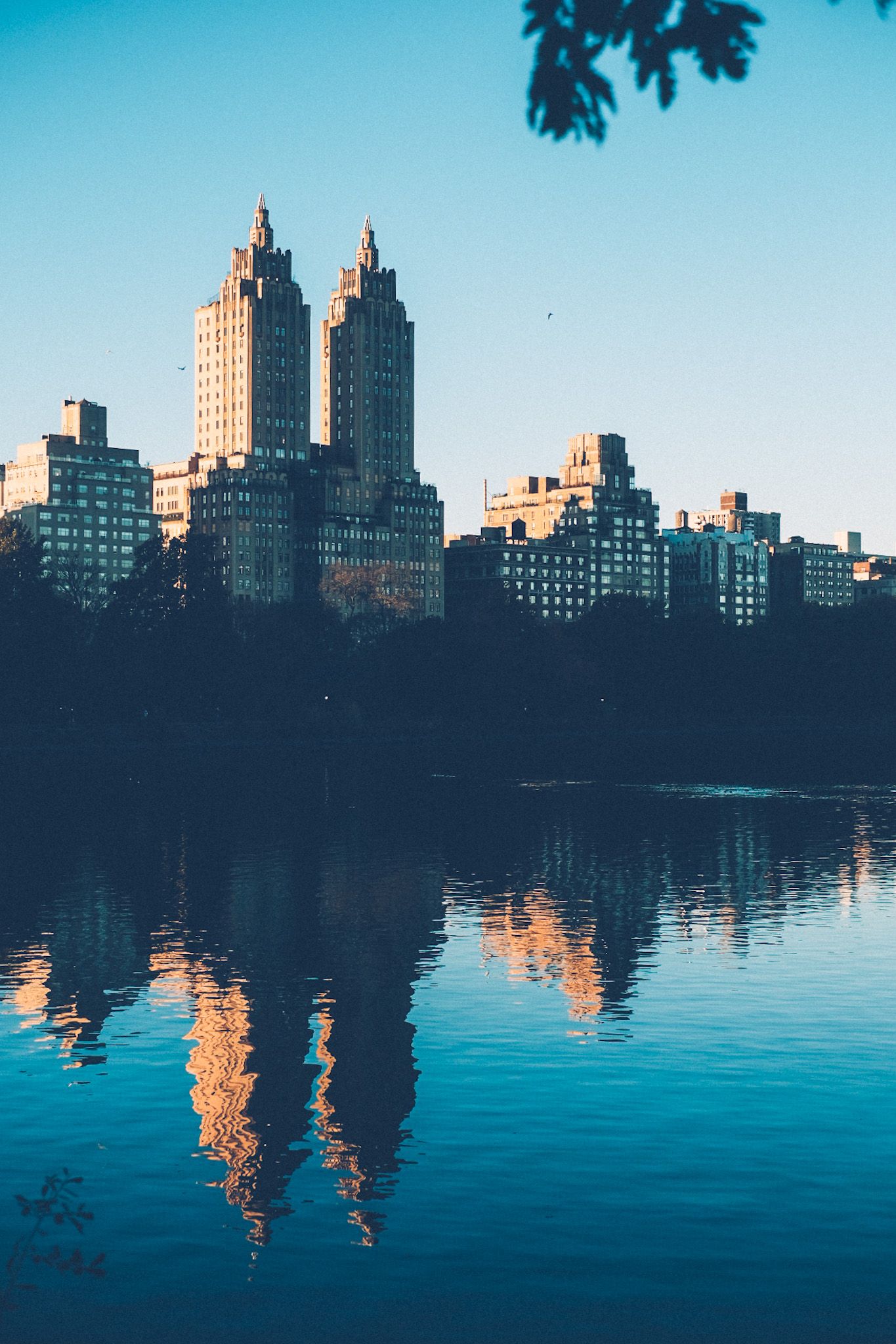 An iconic two-towered building hovers on the edge of Central Park, reflected in the lake on a clear blue-skied day near sunset.