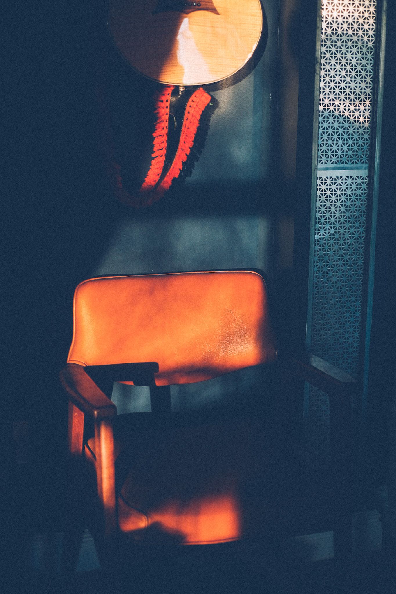 Stripes of afternoon light illuminate the bottom half of a guitar hanging on a wall above an orange leather chair.