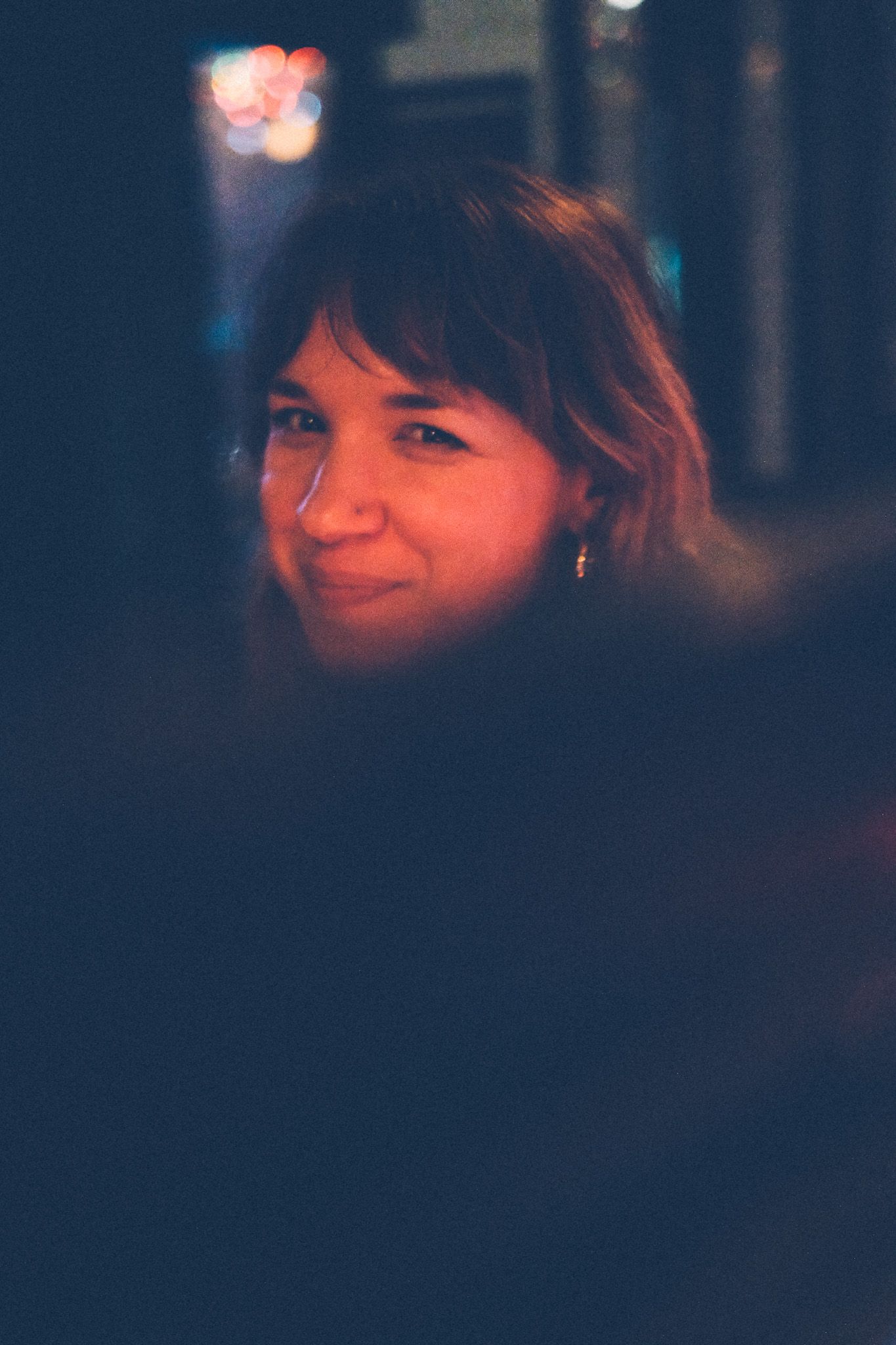 A woman, cast in warm, low light, smiles at the camera. Her lower half below her head is cut off by a dark object in the foreground.