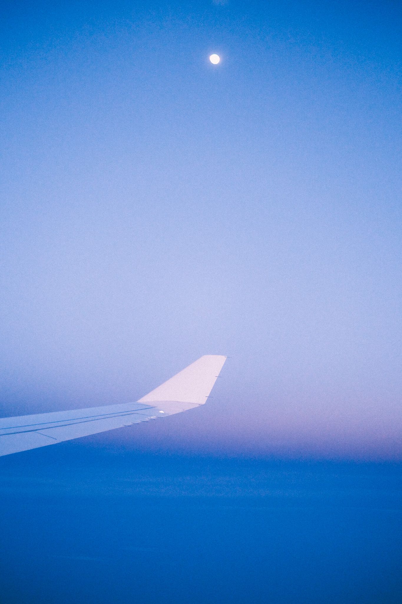The view from the window of a plane at twilight, a purple-blue sky with a moon placed over the wing.
