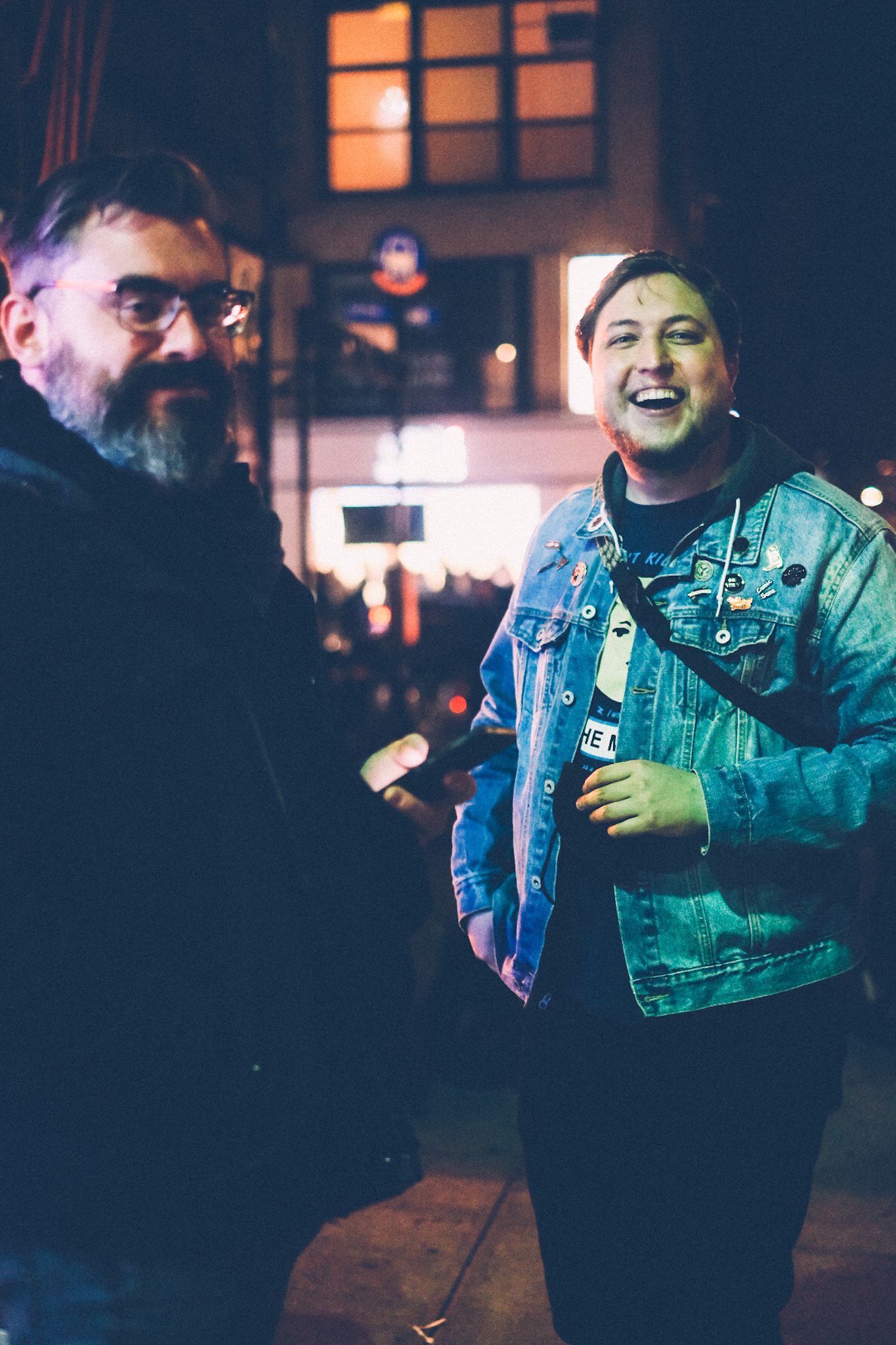 Two people stand outside of a burger joint in the East Village, smiling into the camera, bathed in the neon night.