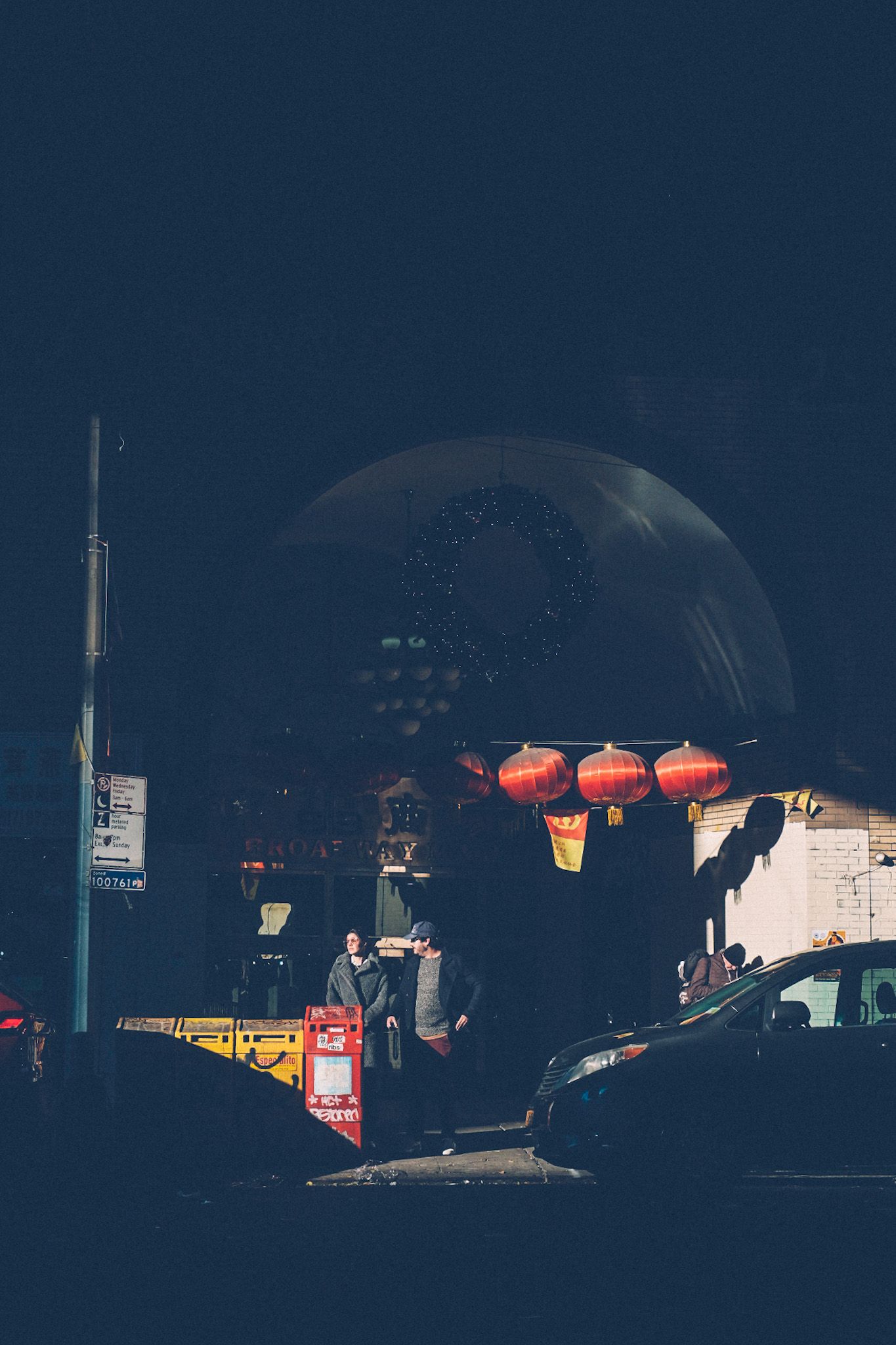 Two people emerge from the East Broadway Mall in Chinatown, New York, mostly cast in shadow with red paper lanterns catching the strip of light.