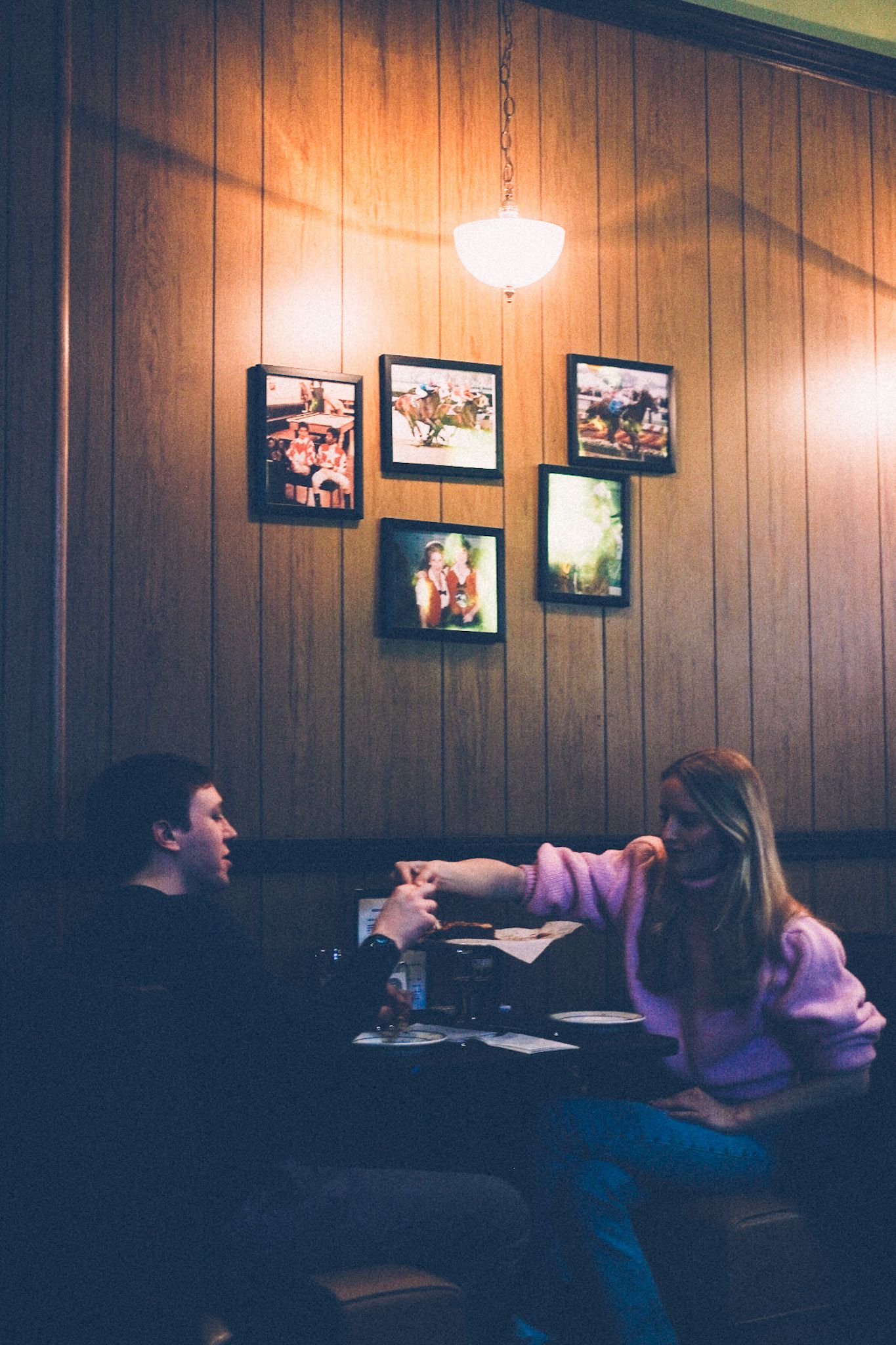 A young couple sits across from each other in a small booth, sharing a pizza. One of them, a woman in a pink sweater, reaches for a slice. Above them hangs a pendant light that illuminates five photo frames on the wood paneled wall above them.