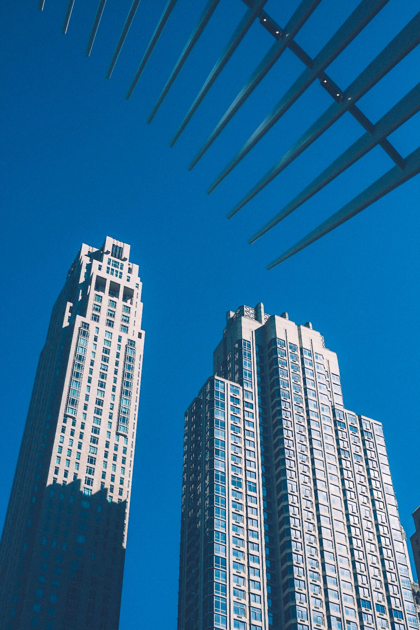 Two buildings rise up from a blue sky while spikes of an unknown origin emerge from the top right corner.