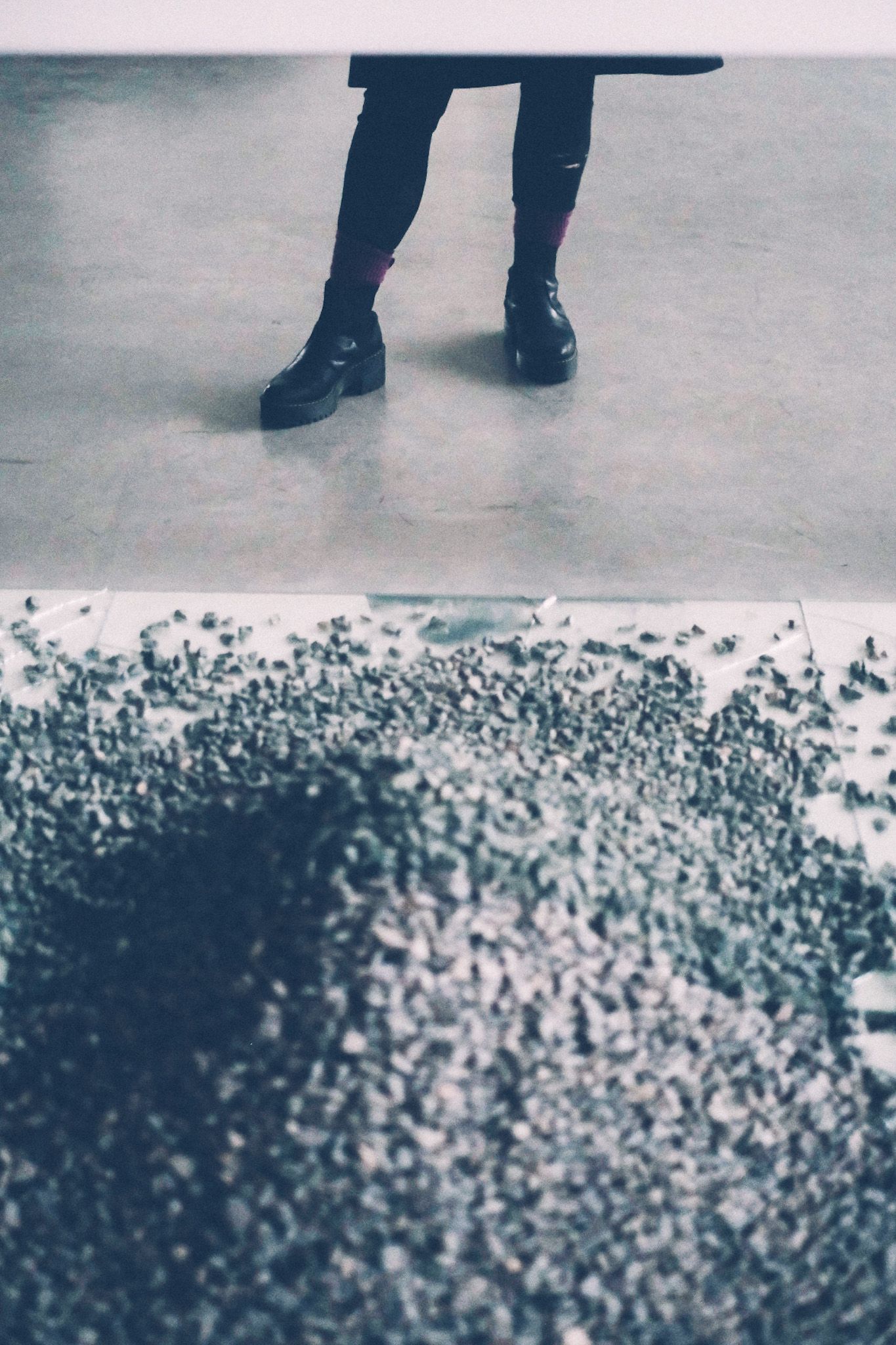 A pair of legs in black boots and tights are reflected in a mirror, standing on polished cement, above apile of gray rocks also reflected in the same mirror.