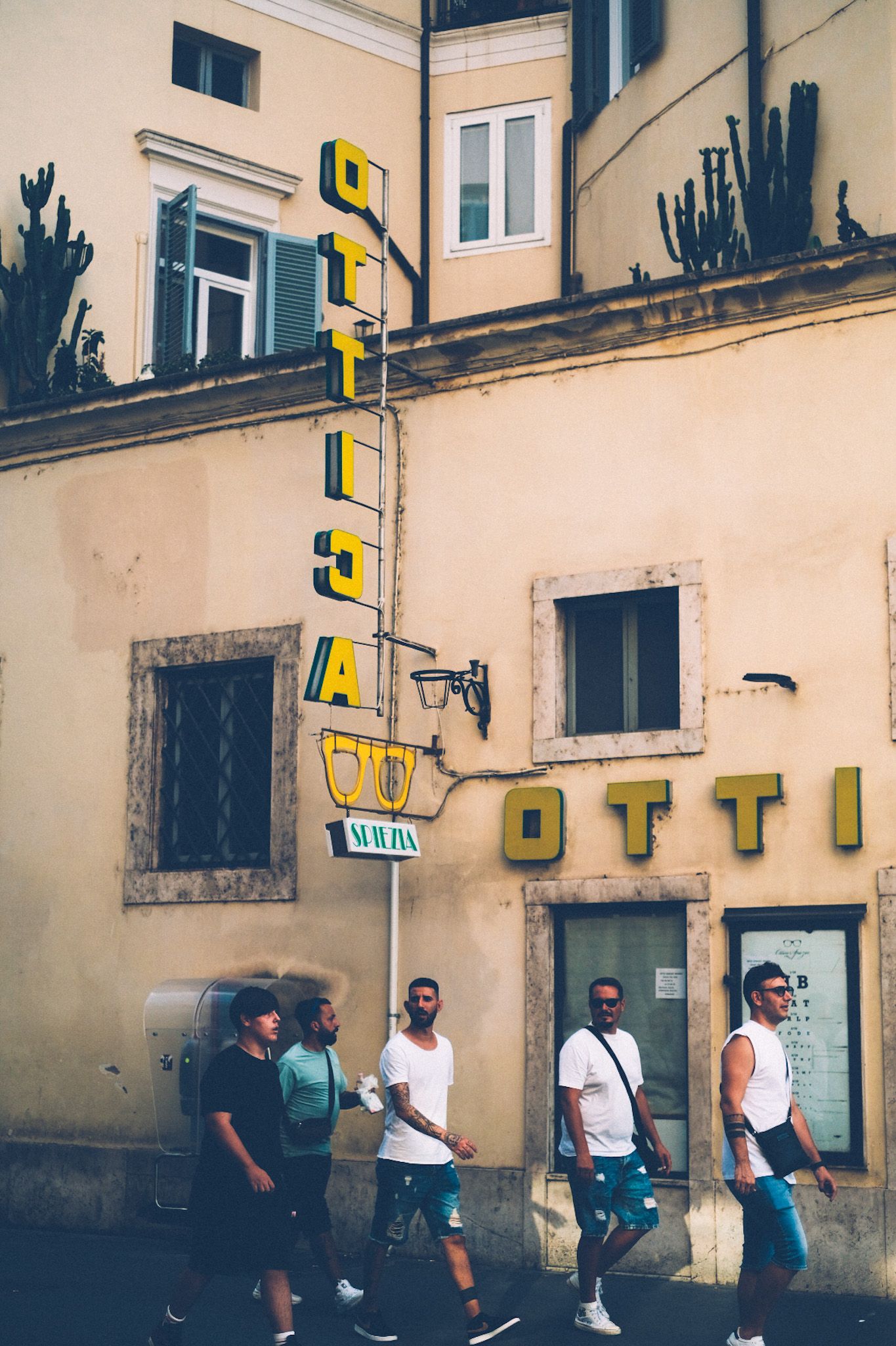 A group of men, mostly in white t-shirts, walks past a yellow sign that says “Ottica” whose bold letters stick out from the building’s faded Roman facade.