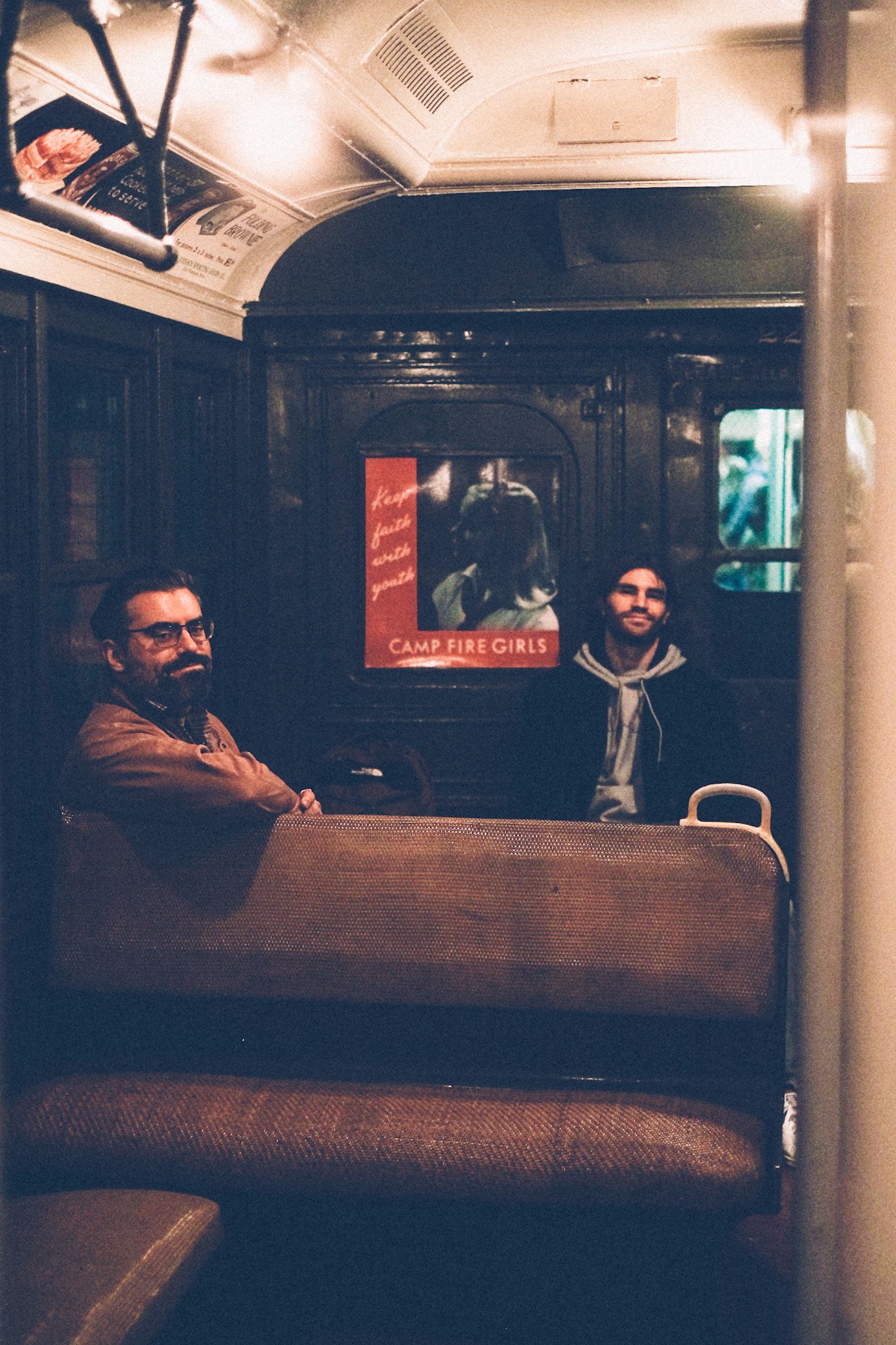 Two men sit in the seats of a vintage subway car in low light.