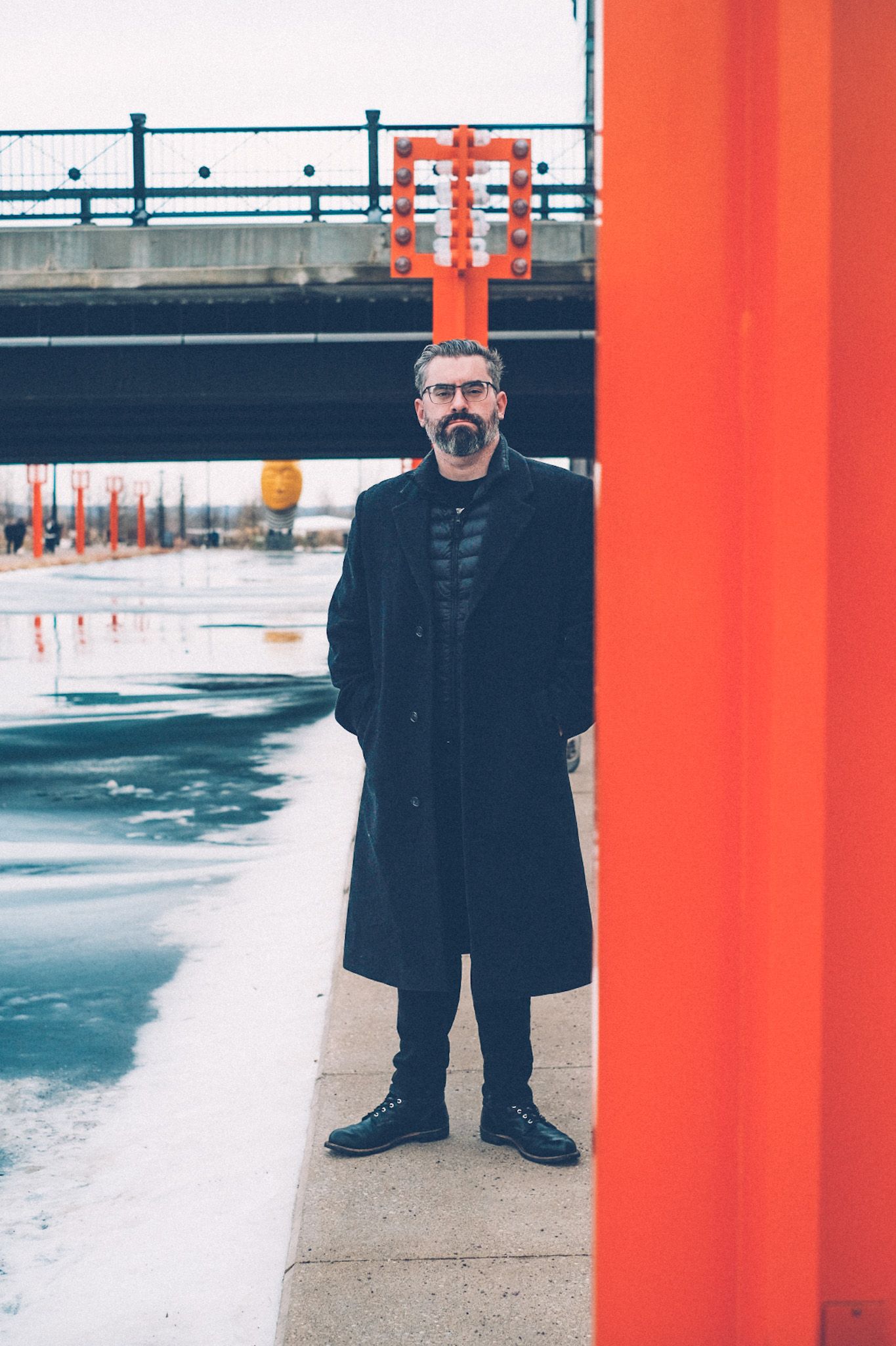 A man in a black peacoat stands beside a frozen body of water next to a sidewalk, partially obscured by an orange lamppost.