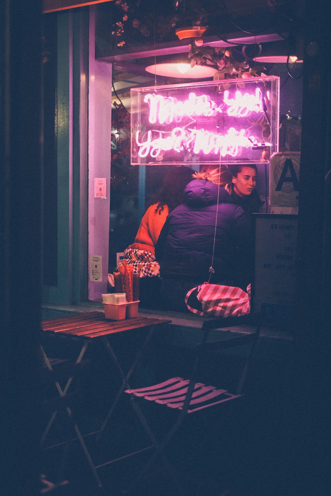In the night, a pink neon sign (letters unreadable because they’re overexposed) illuminates an outdoor sitting area of a restaurant with a slatted wood table and folding chair. Behind the window, inside, a group of women eat.