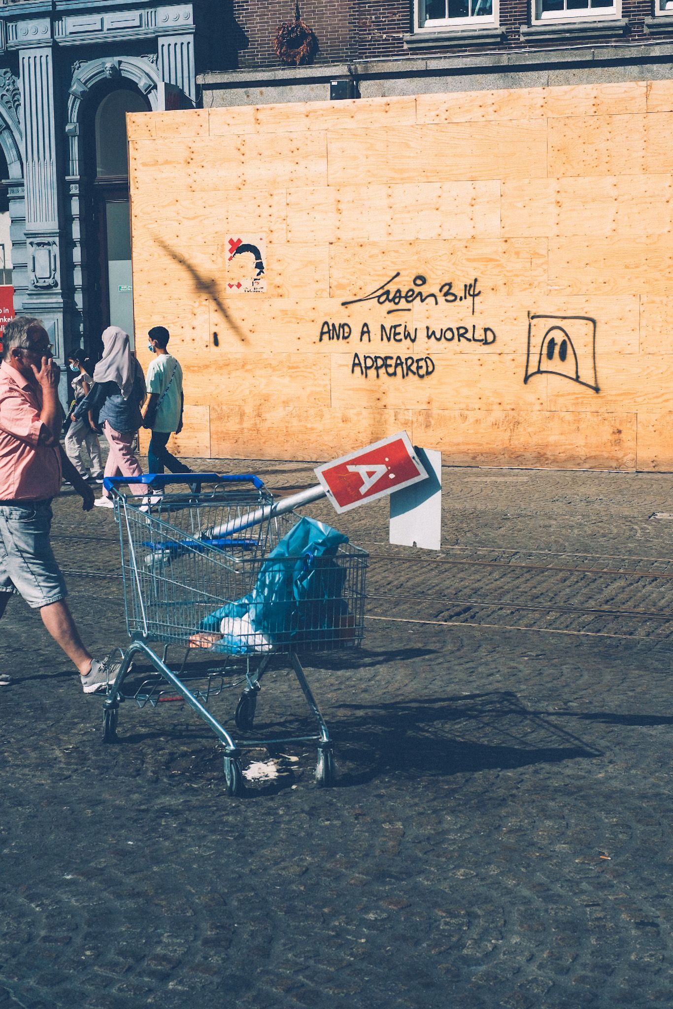 In an open square of Amsterdam, a shopping cart with a little red pennant A hanging off of it stands on the left side of the image. People pass through the sunlit square, in front of a wooden wall that says “And a new world appears” as a bird flies in front of it and leaves a shadow.