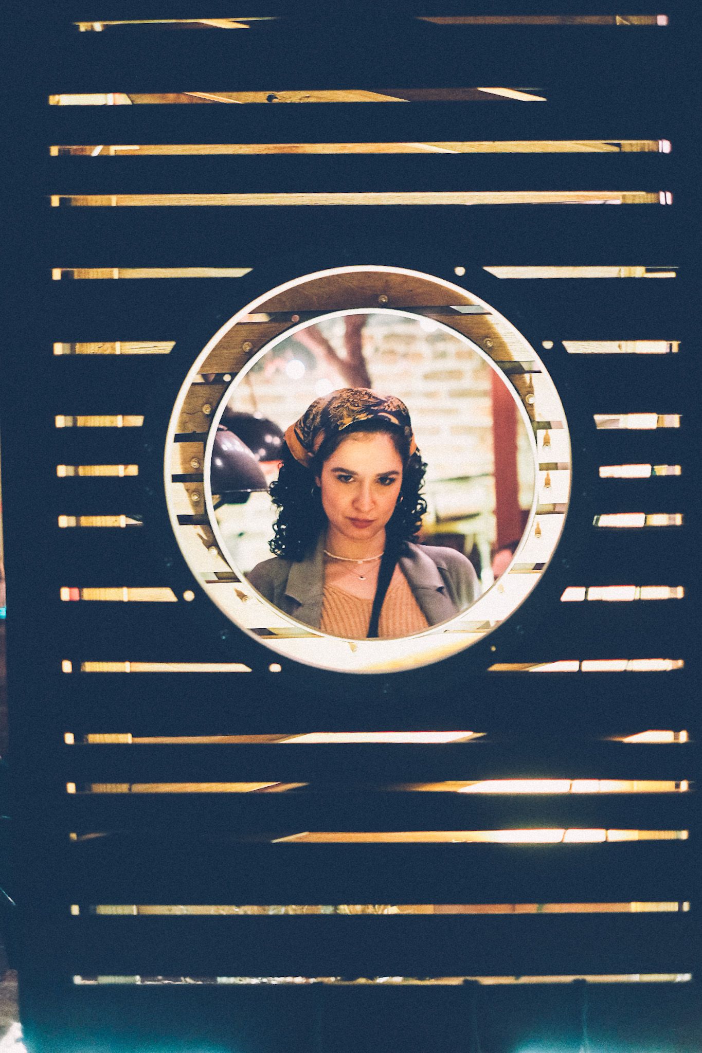 A woman with curly dark hair wearing a bandana looks through a circular cutout in a slatted wood panel, at night in backyard bar.
