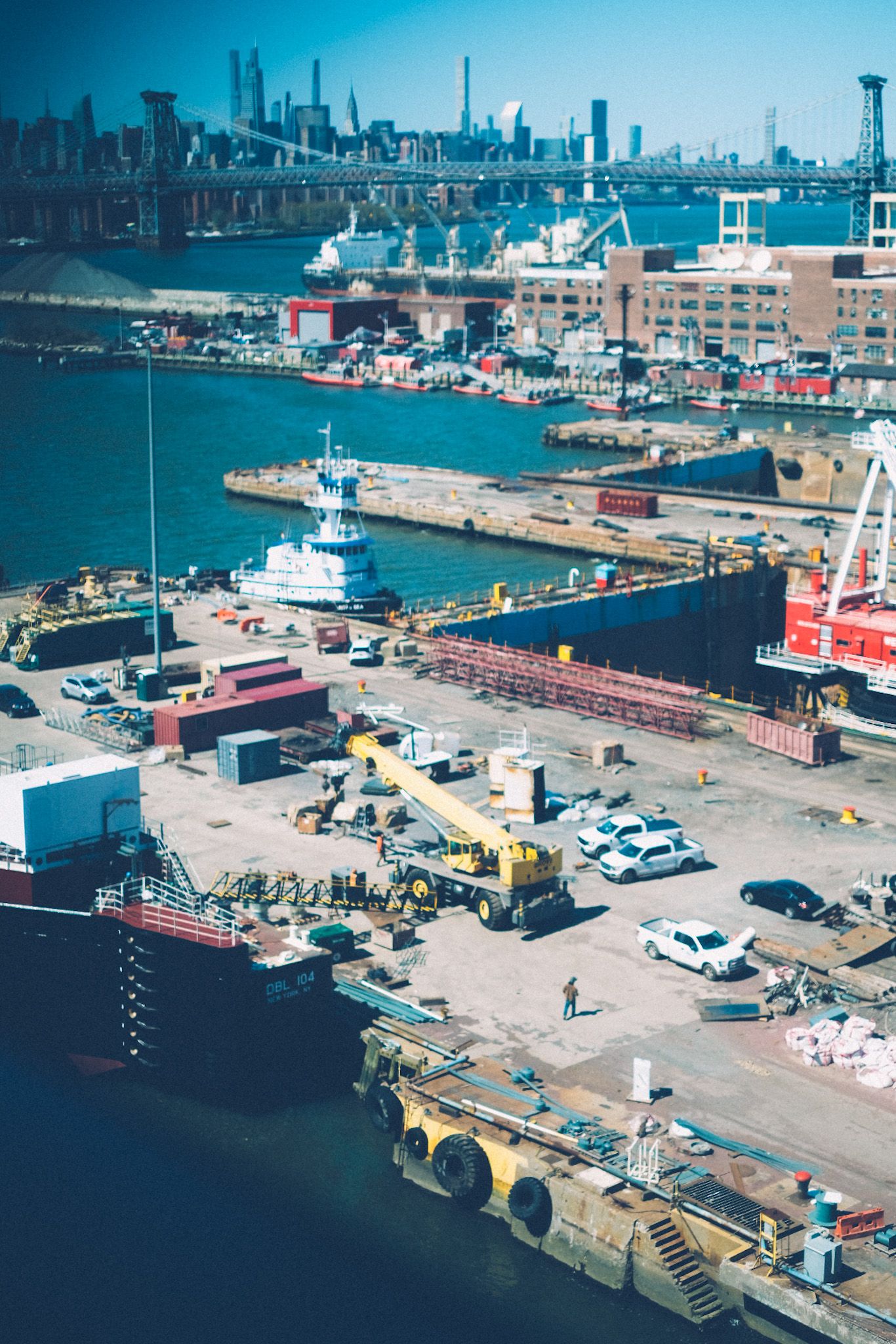 A view outside of a window into the Brooklyn Navy Yard, many construction rigs and boats doing things. The Manhattan skyline peeks up in the background.