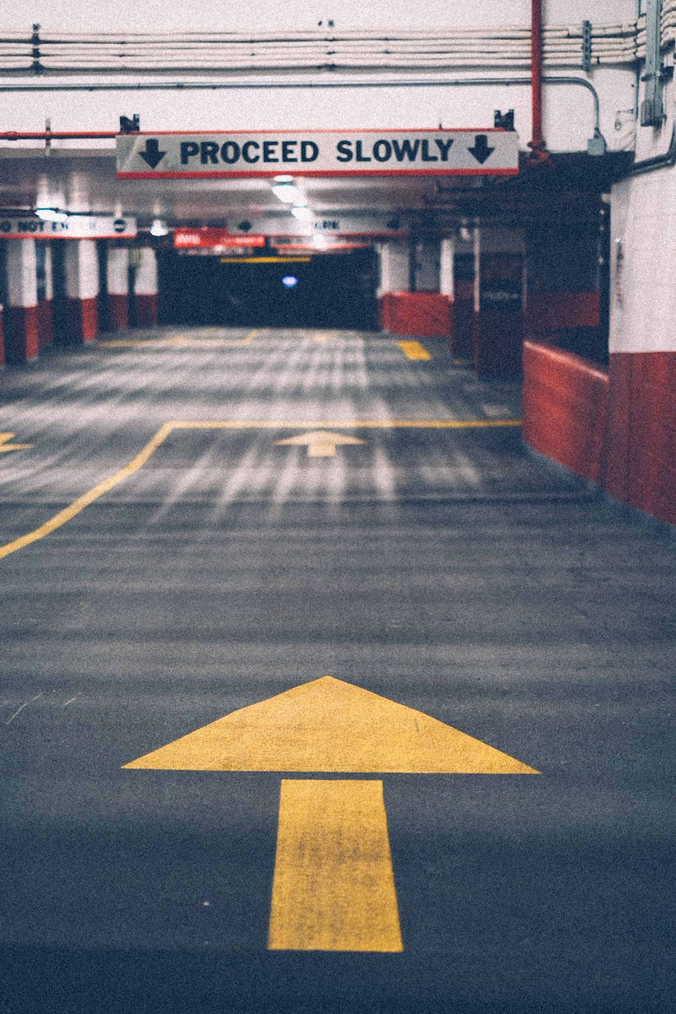 A large yellow arrow painted onto a parking garage floor points inside, continuing into white and red walls and gray cement and eventually into the distant dark. A sign above says “Proceed slowly.”