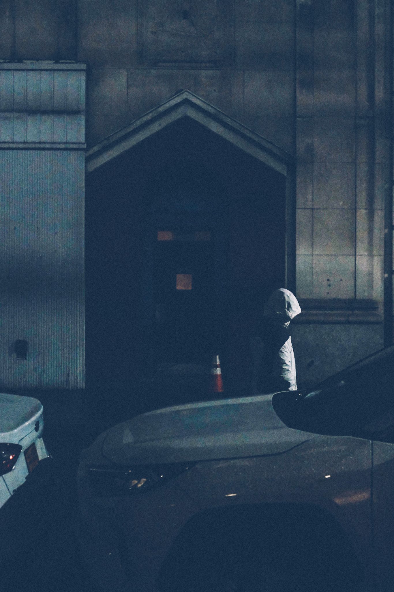 At night, a hooded figure walks past a darkened doorway.