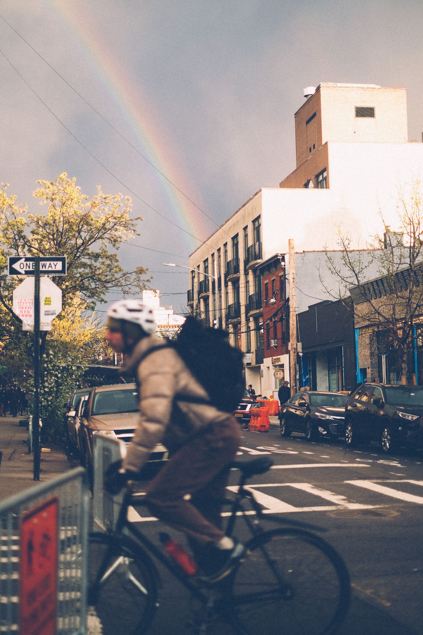 The sun sets in a gray sky on a street in Brooklyn, revealing the arch of a rainbow. In the foreground, out of focus, a man rides past on a bike.