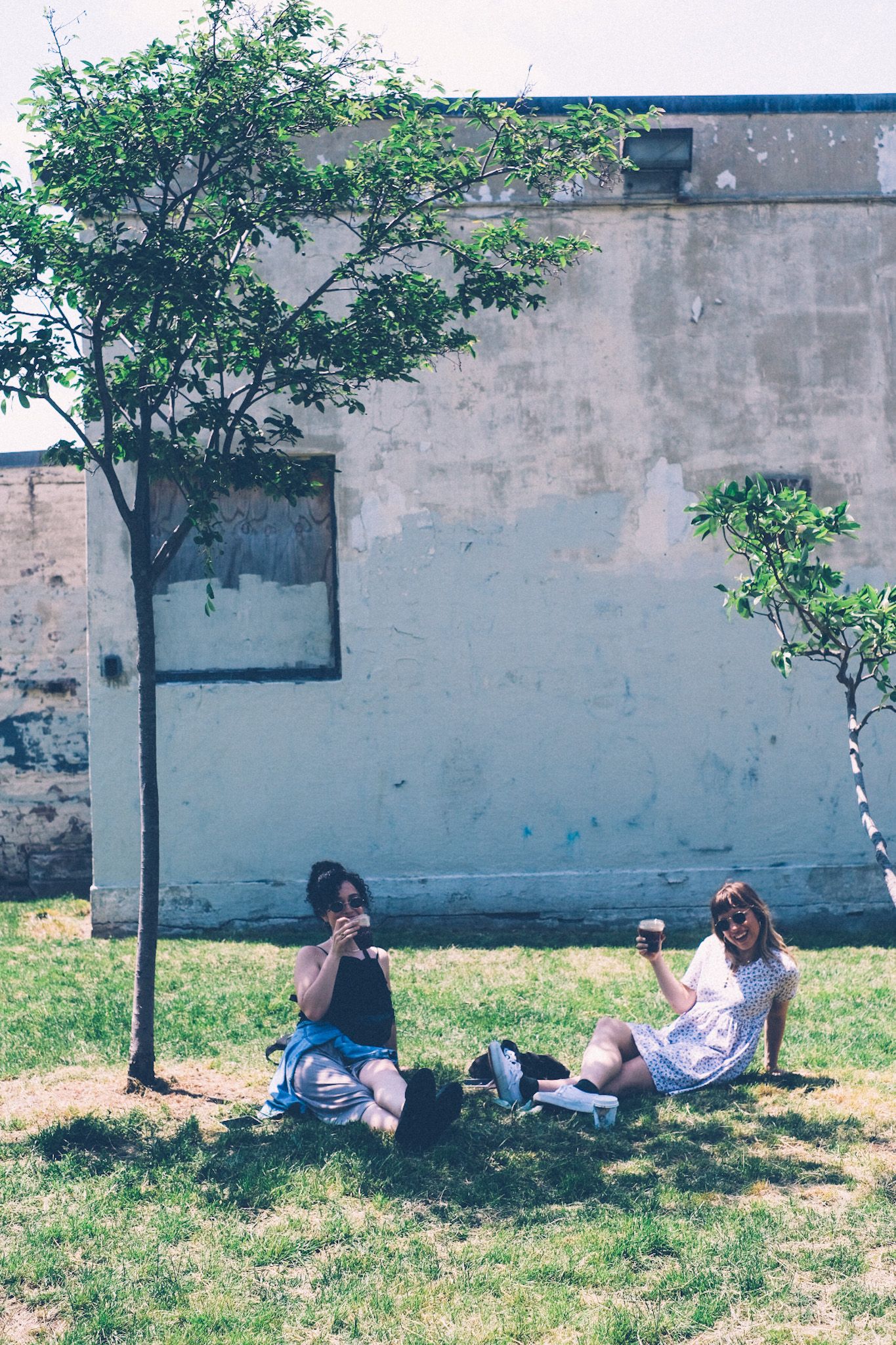 Two women sit in the grass, under small trees, in front of what looks to be an abandoned building. They are smiling.