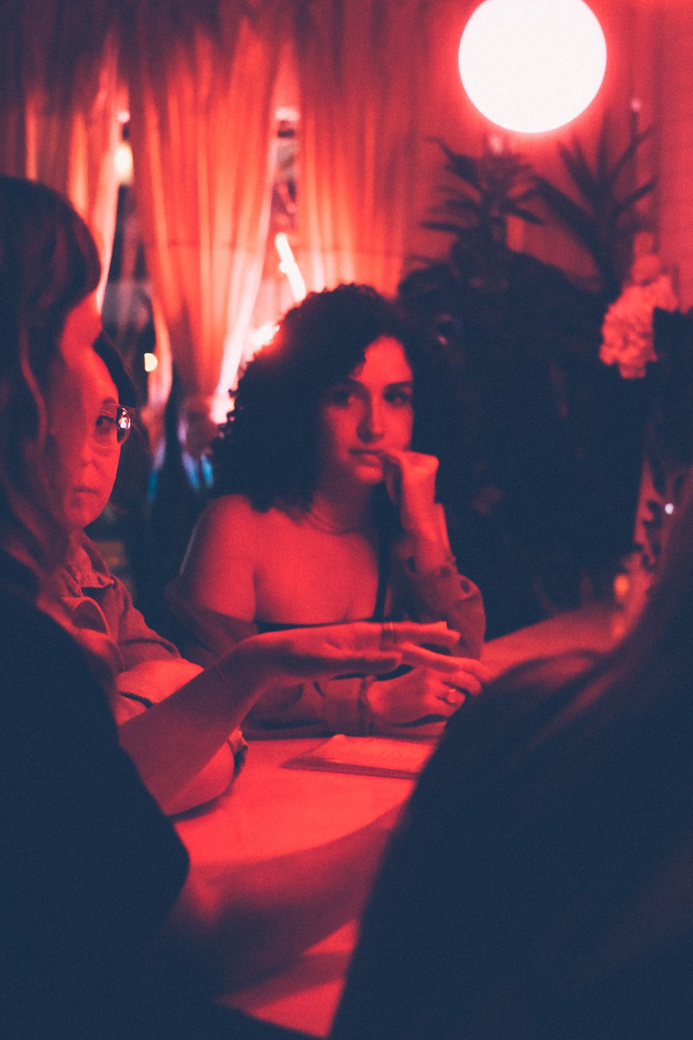 A woman sits in a bar among friends, bathed in red light, looking like something is on her mind.