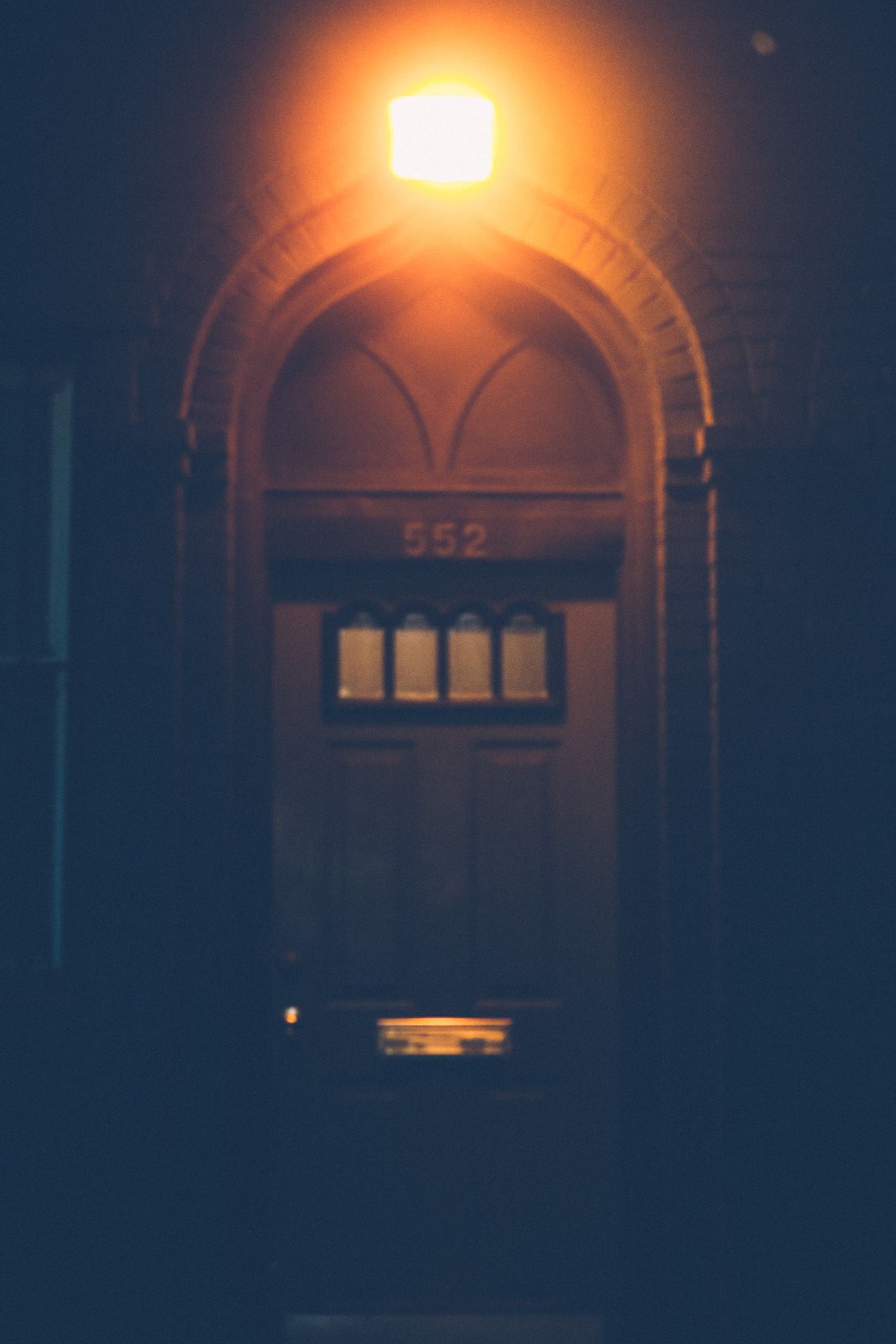 A front door at night is just slightly out of focus, bathed in an orange light right above the arched door frame with the number 552 underneath it.