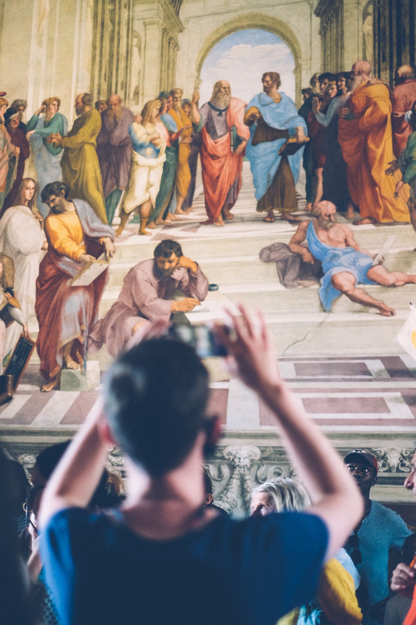 In the foreground out of focus, a man holds up a phone to take a photo of Raphael’s painting “The School of Athens” in the Vatican.