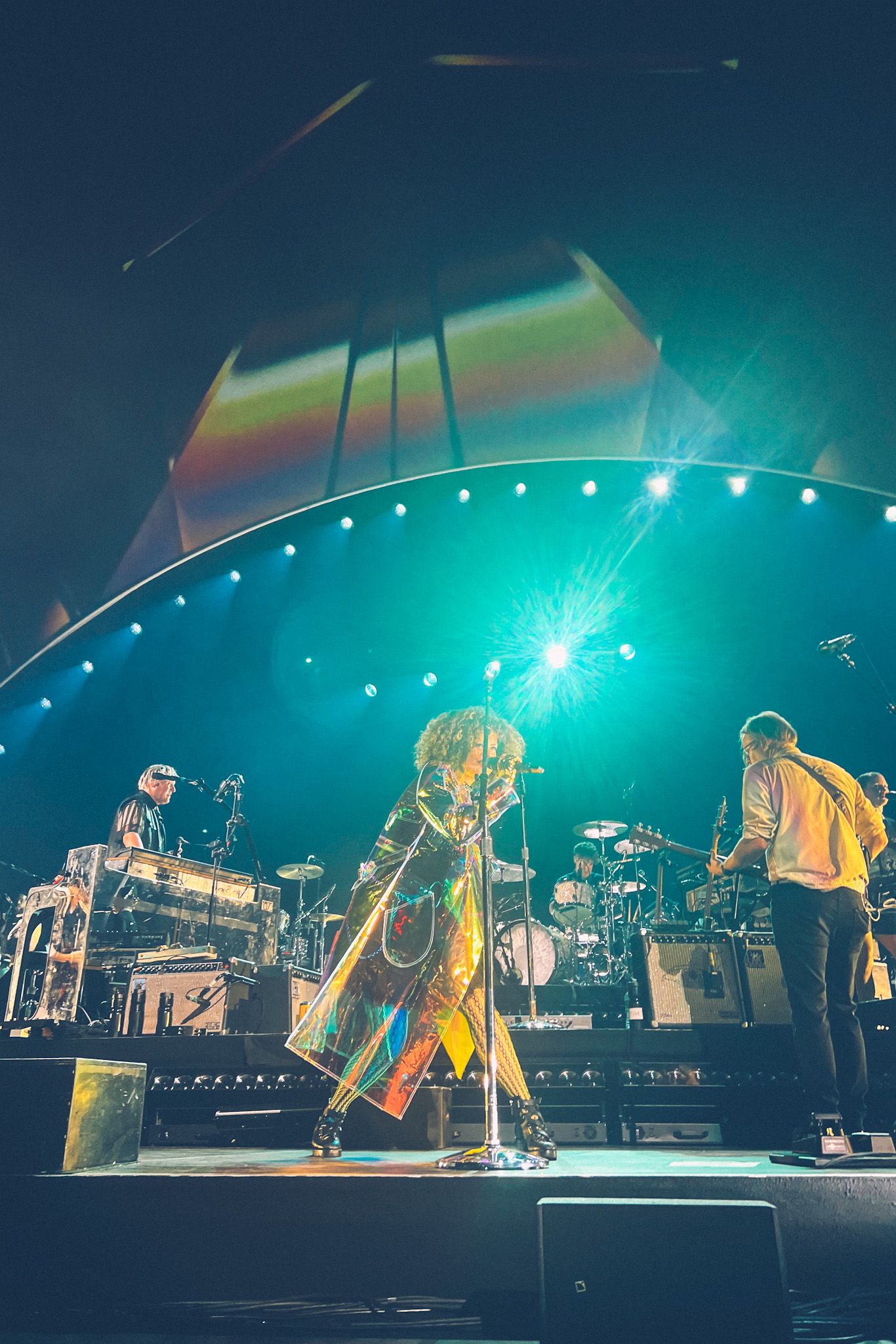 The front woman of the band Arcade Fire struts across a stage wearing a reflective rainbow raincoat, the band in the background alongside blue and rainbow lighting.