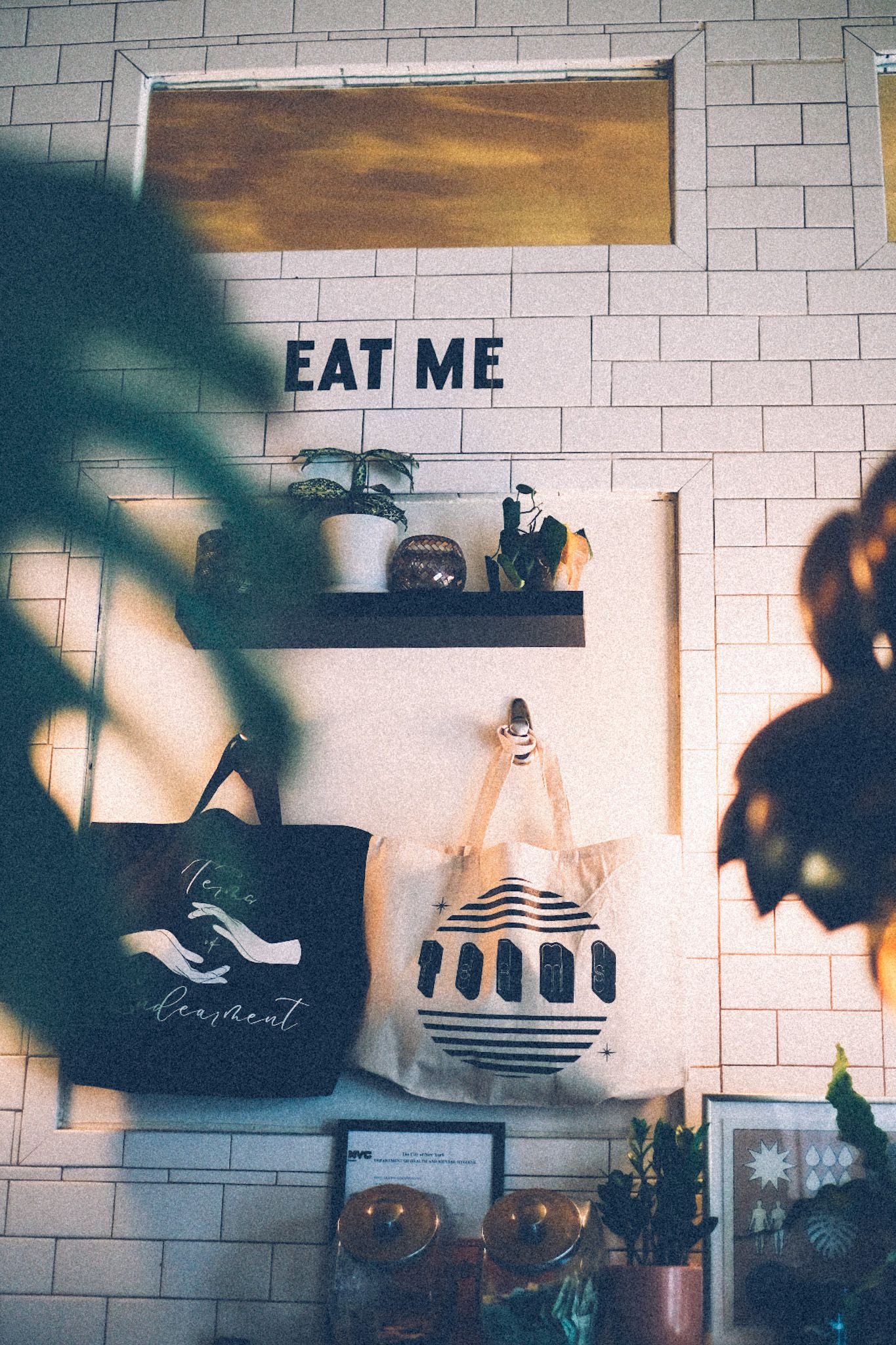 The words “eat me” are adhered against white brick in a coffee shop, framed by hip tote bags and plants half-obscuring the photo.
