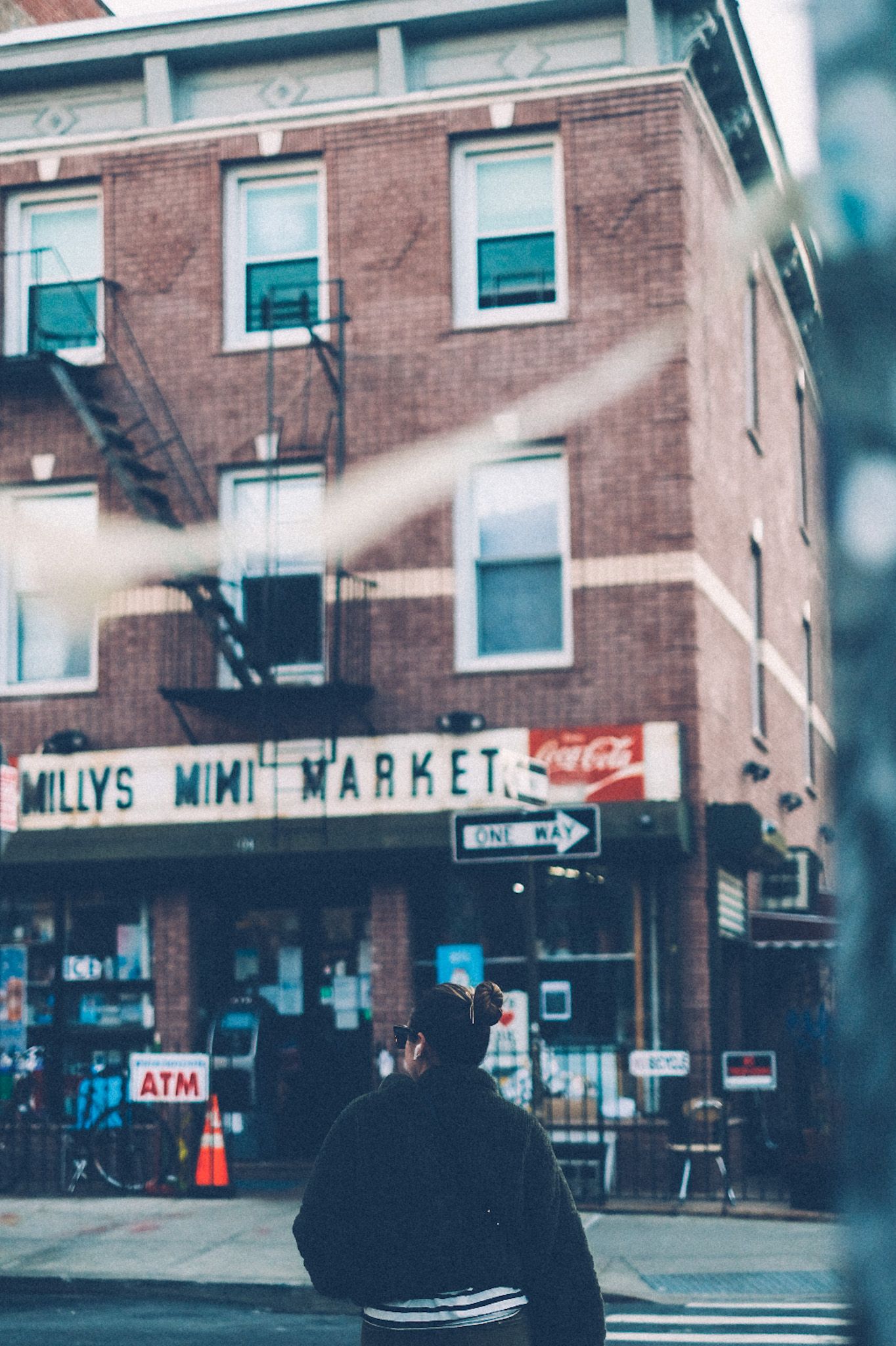 From across the street, a sign on a three-story brick building says Milly’s Mini Mart. A woman is crossing the street and a piece of tape from the telephone pole blows across the foreground.
