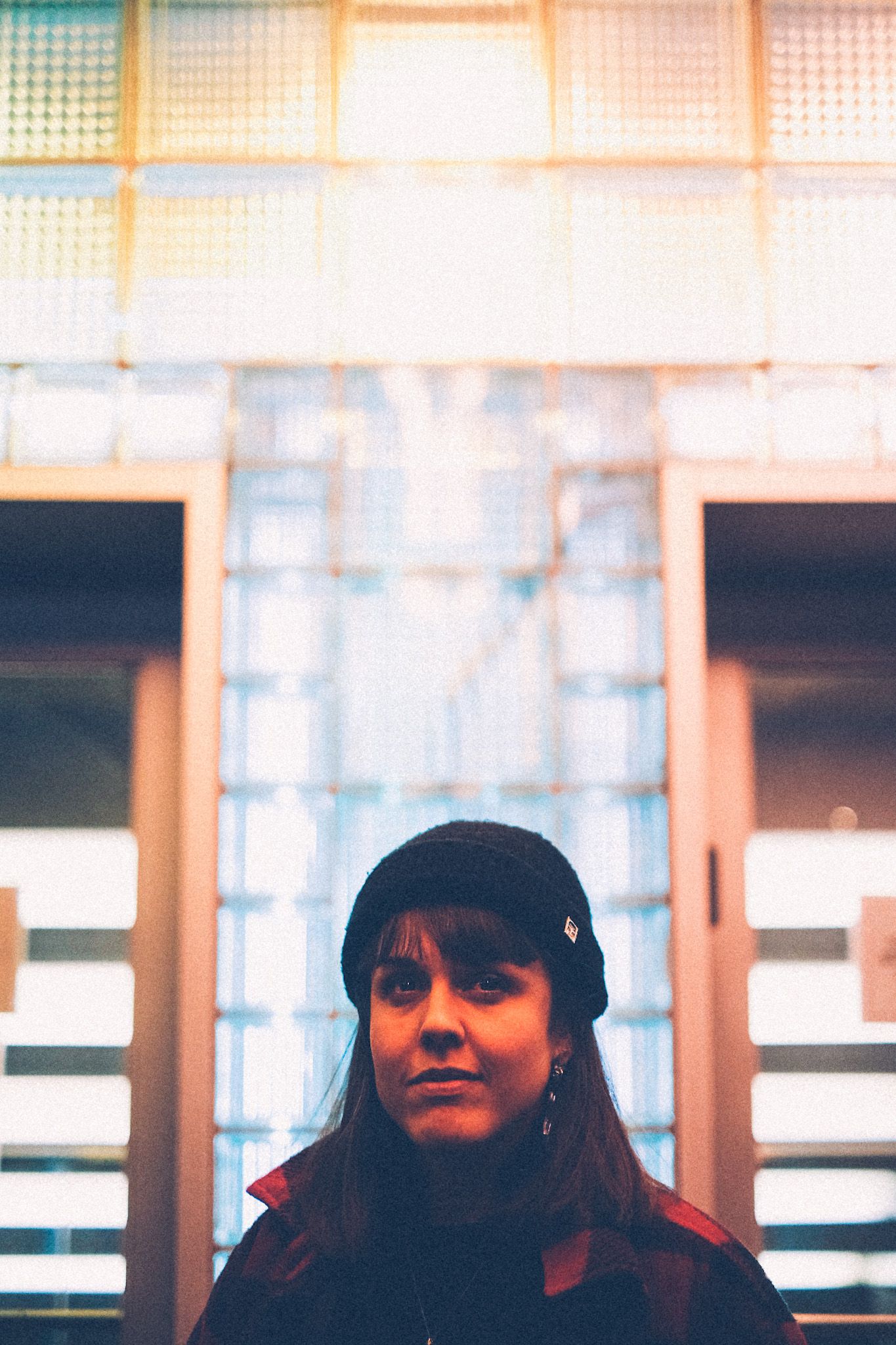 A woman in a black beanie looks up, under the lights of a theatre awning. In the background are squares of frosted white-blue glass between two doors.