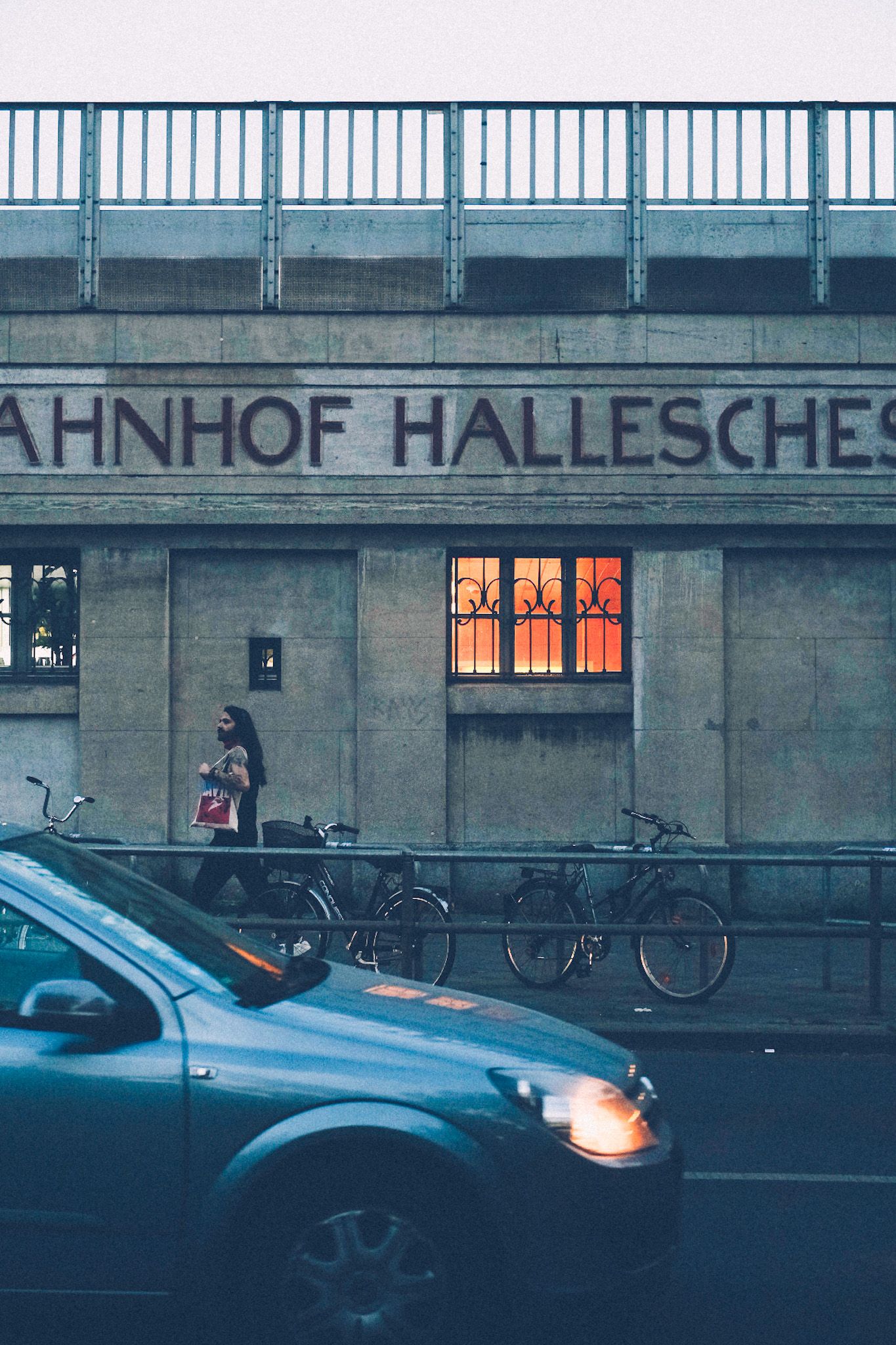 In the light evening, a woman passes by a building with the words “Bahnhof Hallesches” on it, slightly cropped. A square window in the middle of the picture emanates orange light through decorative iron bars.
