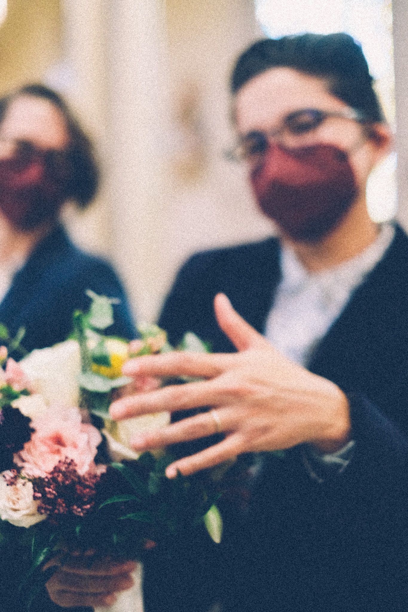 Two women in well-tailored blue suits and maroon masks lean against church pews, out of focus. The one in the foreground flashes her hand over a bouquet of burgundy, pink, and white wedding flowers.