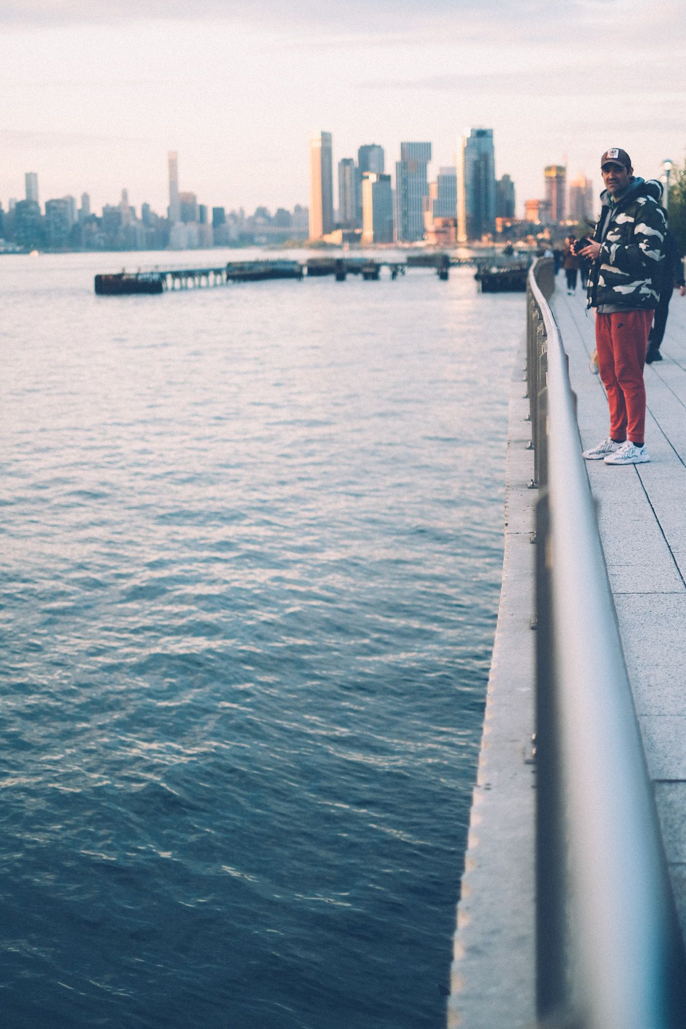 A man in red pants is at the very right edge of the frame, next to a long railing, the skyline of north Brooklyn in the background near sunset on the river.