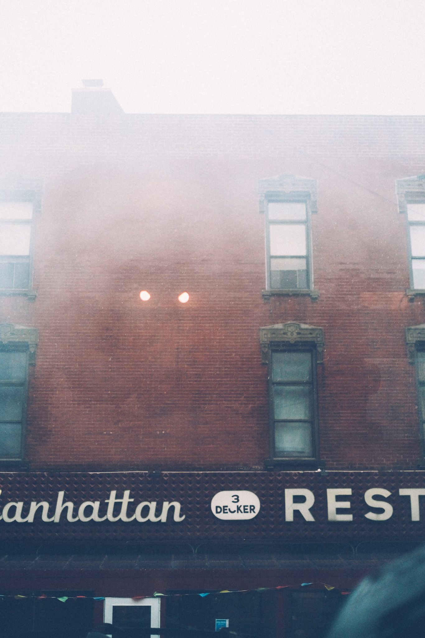 In the fog, a brick building ascends into the cloudy sky above, with the cutoff words “Manhattan Restaurant” and “3 decker” on the bottom.