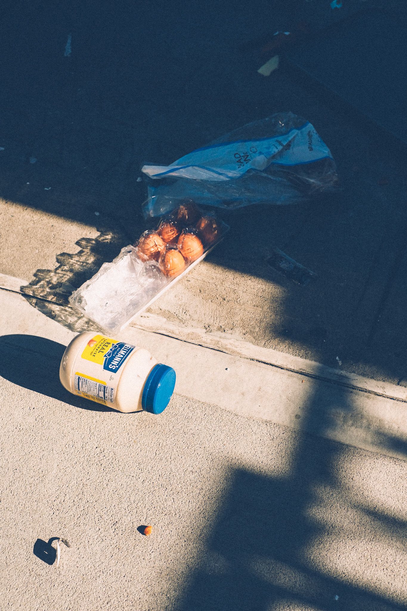 A jar of mayonnaise, a half-full carton of eggs and an empty bread bag lie splayed on the sidewalk in the stark light and shadow of the afternoon.