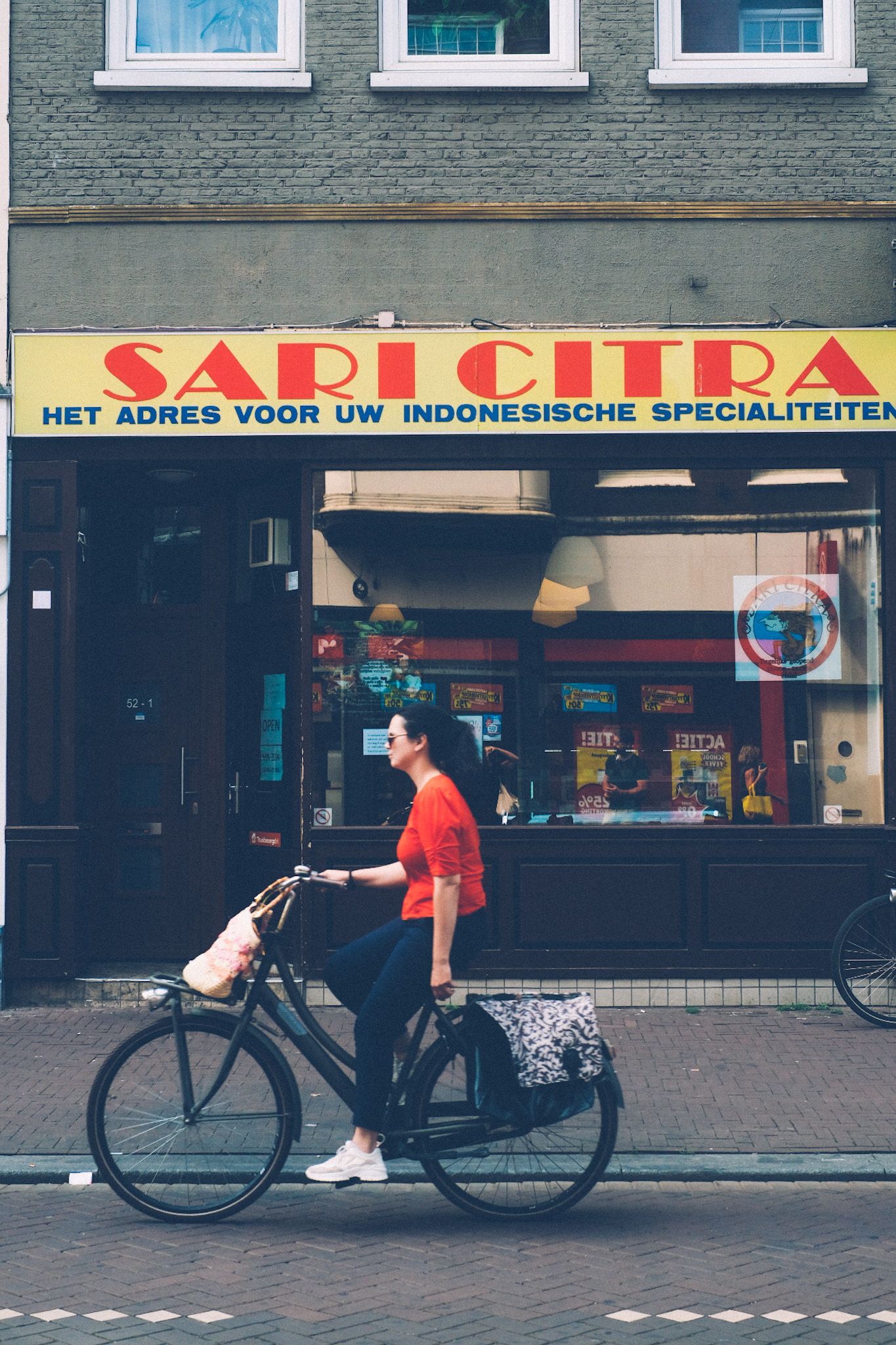 A woman in a red shirt on a bike passes in front of a building, a sign with bright red letters to match her shirt saying “Sari Sitra.”