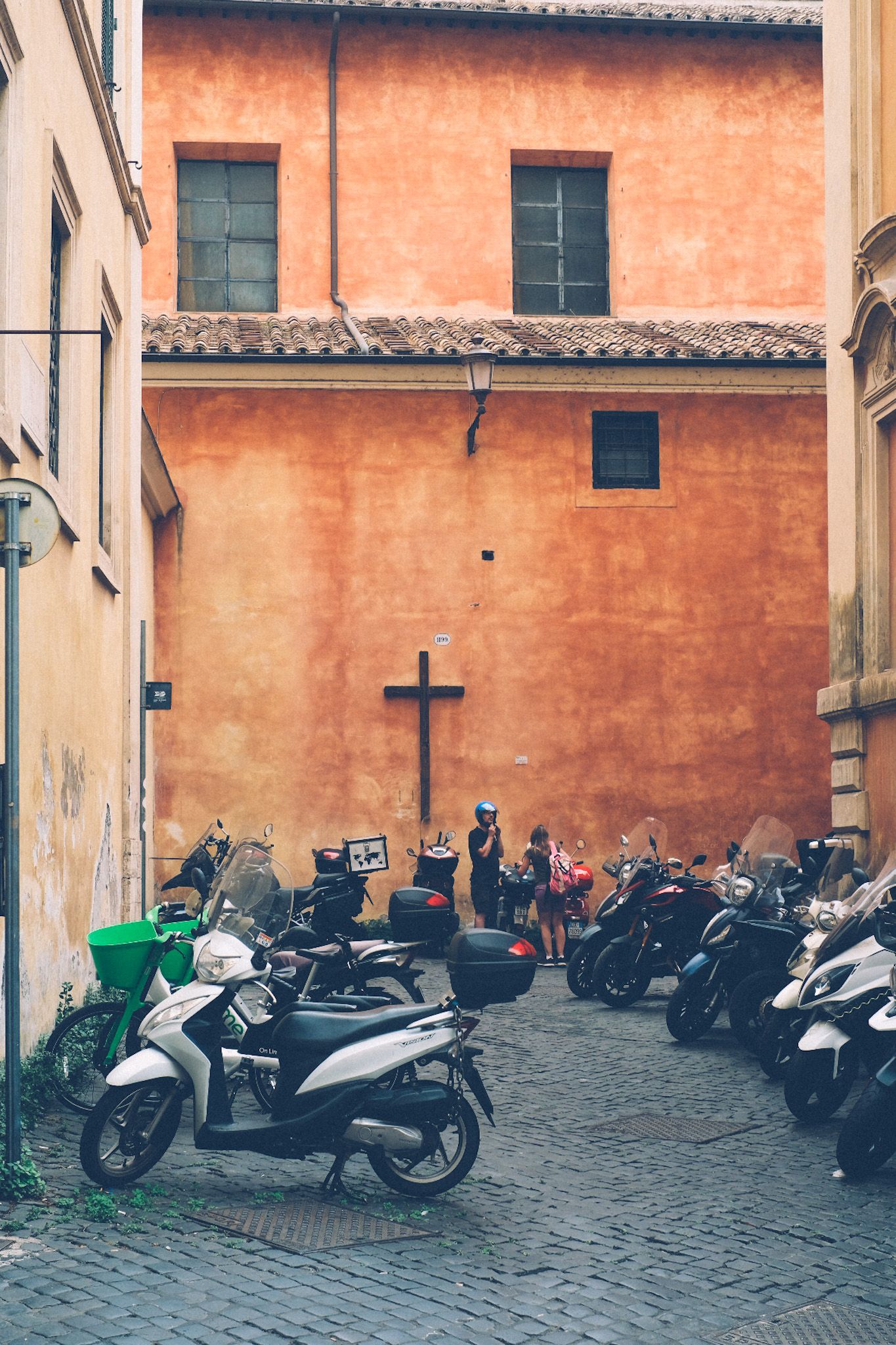 A wooden cross hangs outside on an orange wall of a building, a small square filled with motorcycles.