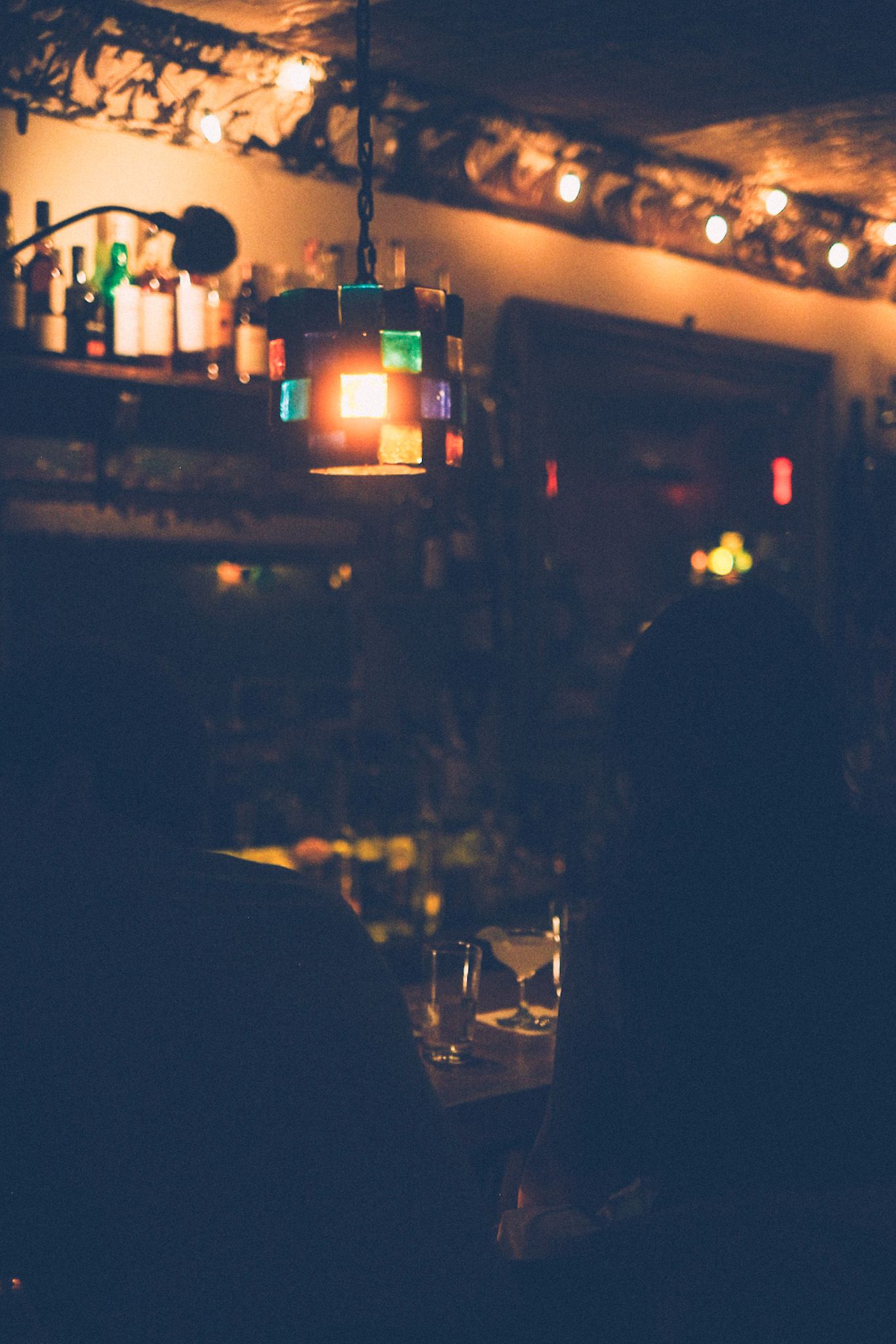 Two silhouetted sit at a dimly lit bar, booze bottles and crown molding and a multicolored stained glass lamp all crowded in the background.