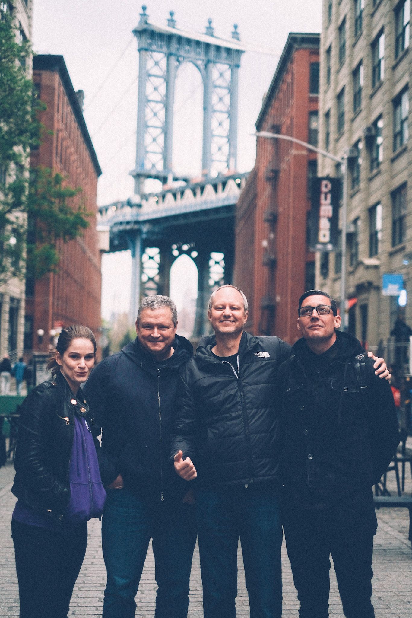 Four people stand smiling in front of the iconic view of the Manhattan bridge from Brooklyn.
