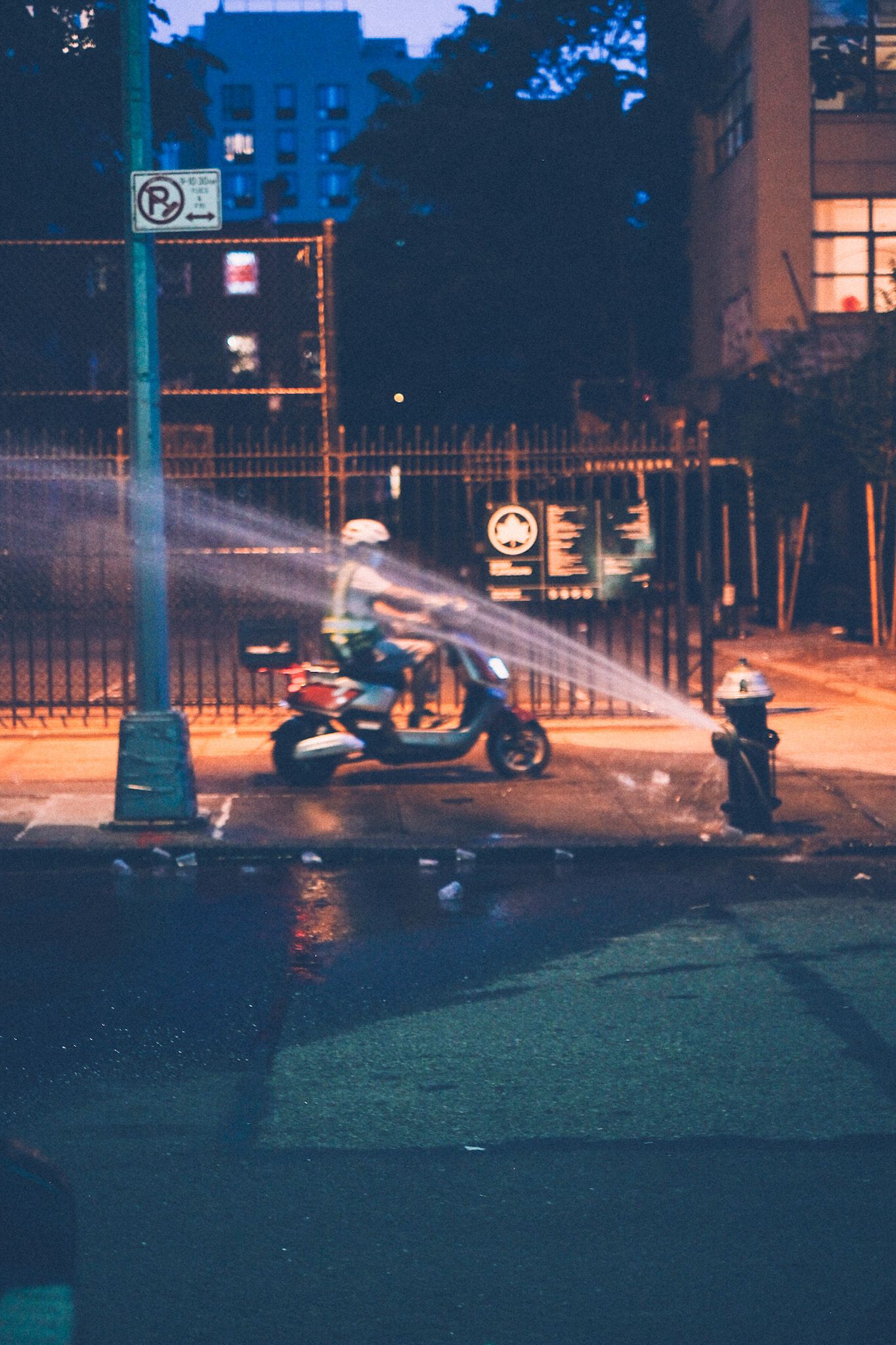 A fire hydrant is set loose on a city street at night, lit with golden street lamp. A man on a motorcycle rides past in a blur.