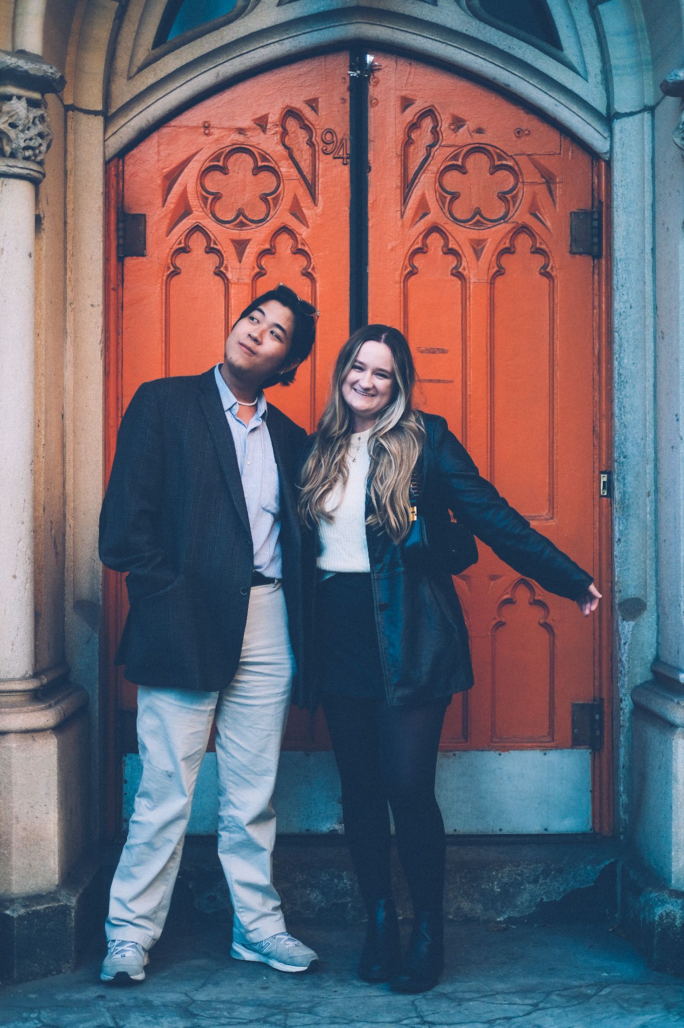 Two people stand smiling in front of a bright orange door of what looks to be a church, decorated with woodcut patterns.
