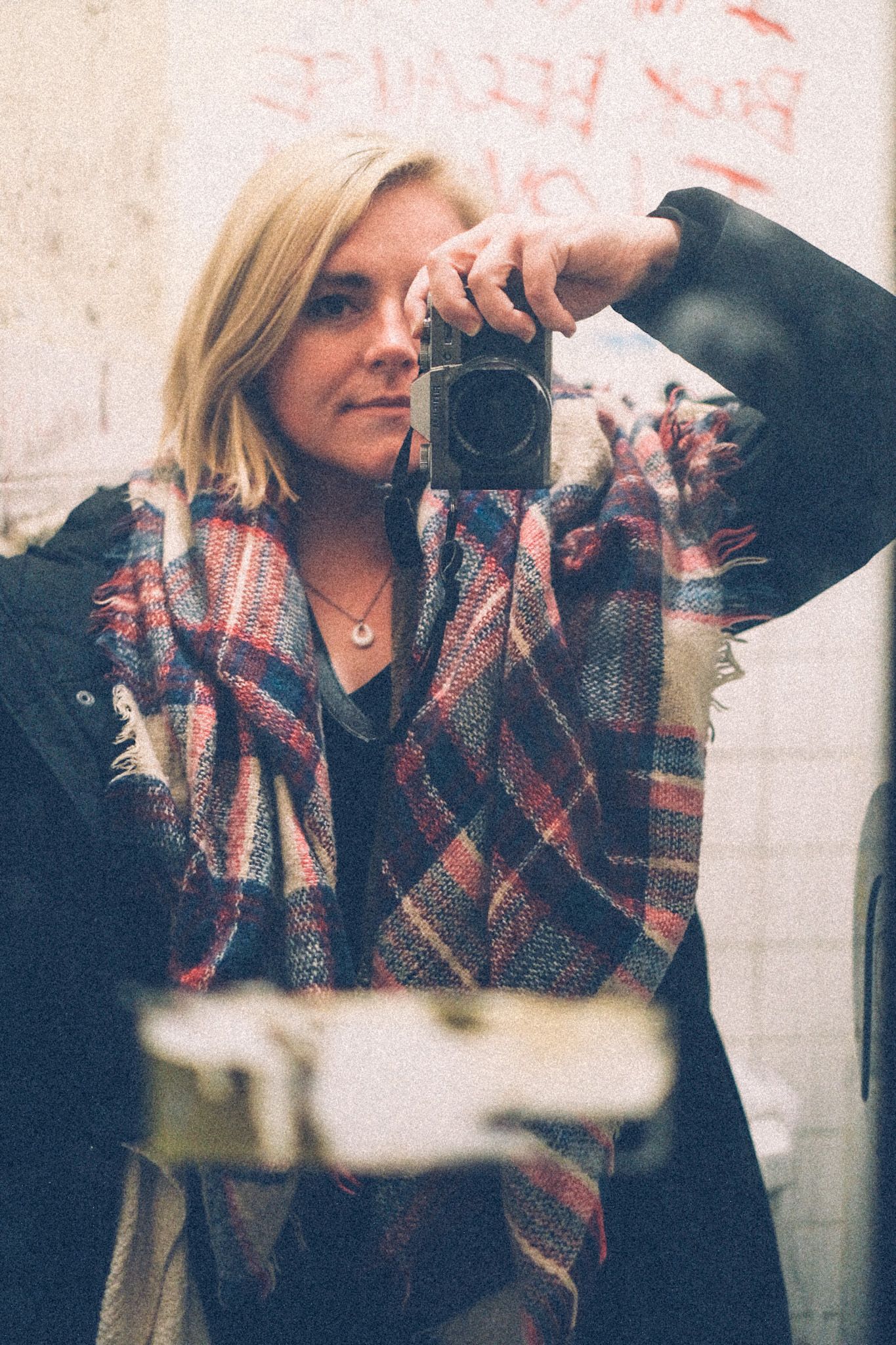 A self-portrait of a woman in a mirror hung on a dive bar bathroom. She is blond with a pink patterned scarf. The camera is out of focus in the reflection.